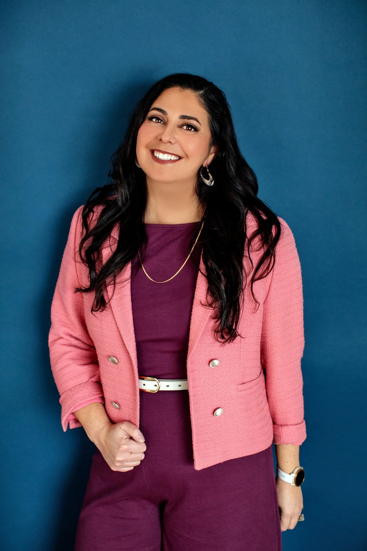 A woman with long black hair and a bright smile stands against a blue wall. She is wearing a pink blazer, a purple dress, a white belt, and accessorizes with earrings, a necklace, a watch, and rings.