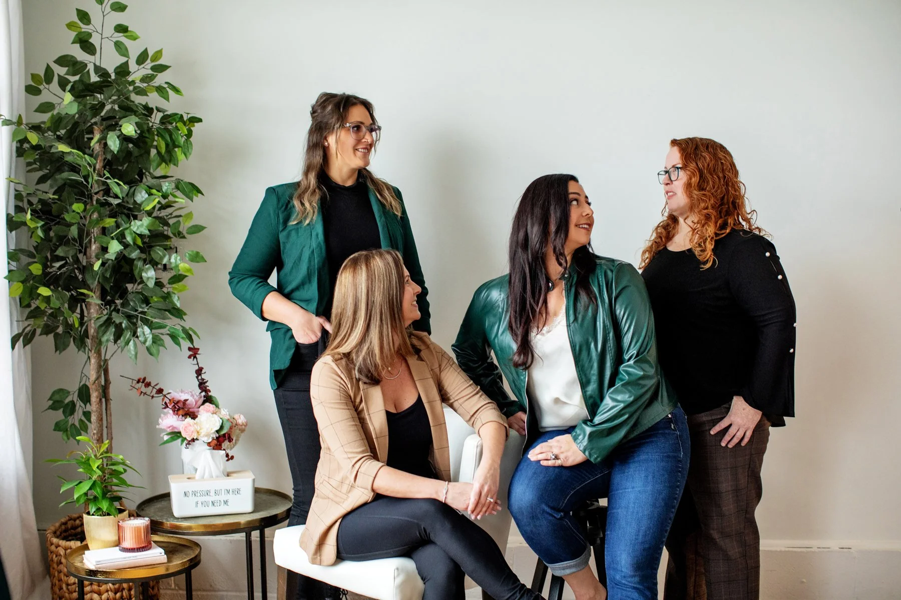 Five women having a conversation in a room with a tall potted plant, a side table with flowers, and a neutral wall background.