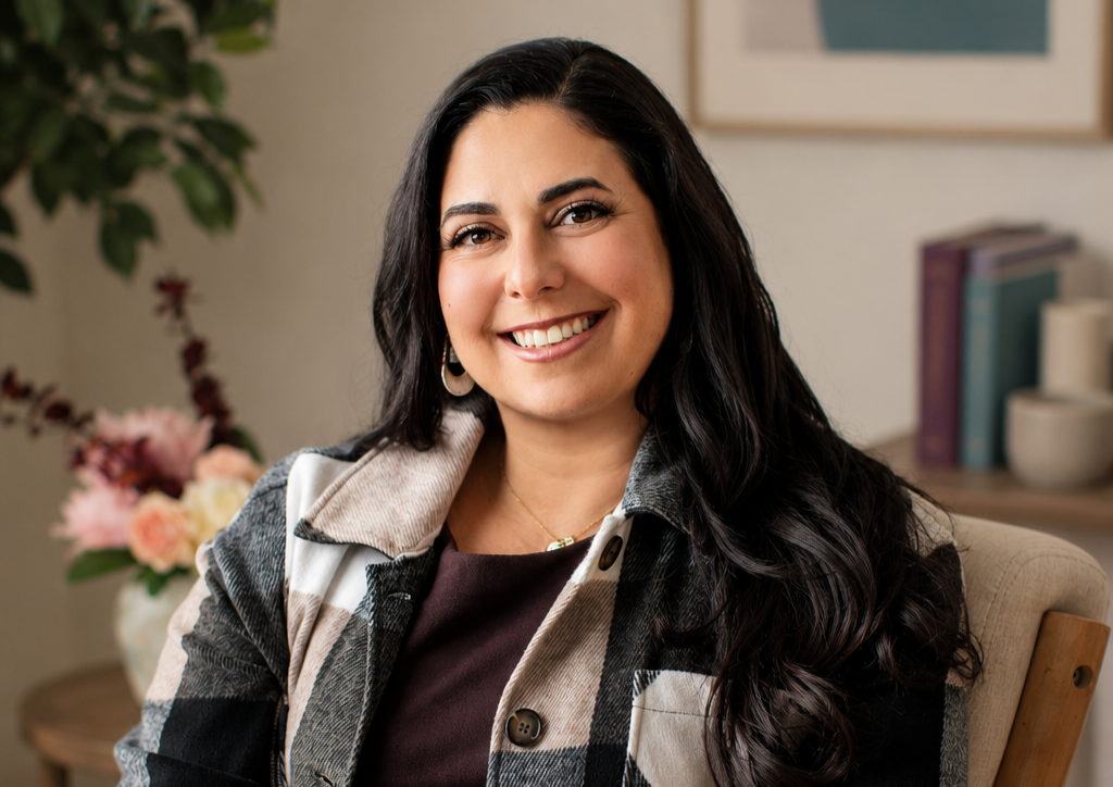 A woman with long dark hair smiling at the camera, sitting in a cozy indoor setting with a plant and a bouquet of flowers in the background.