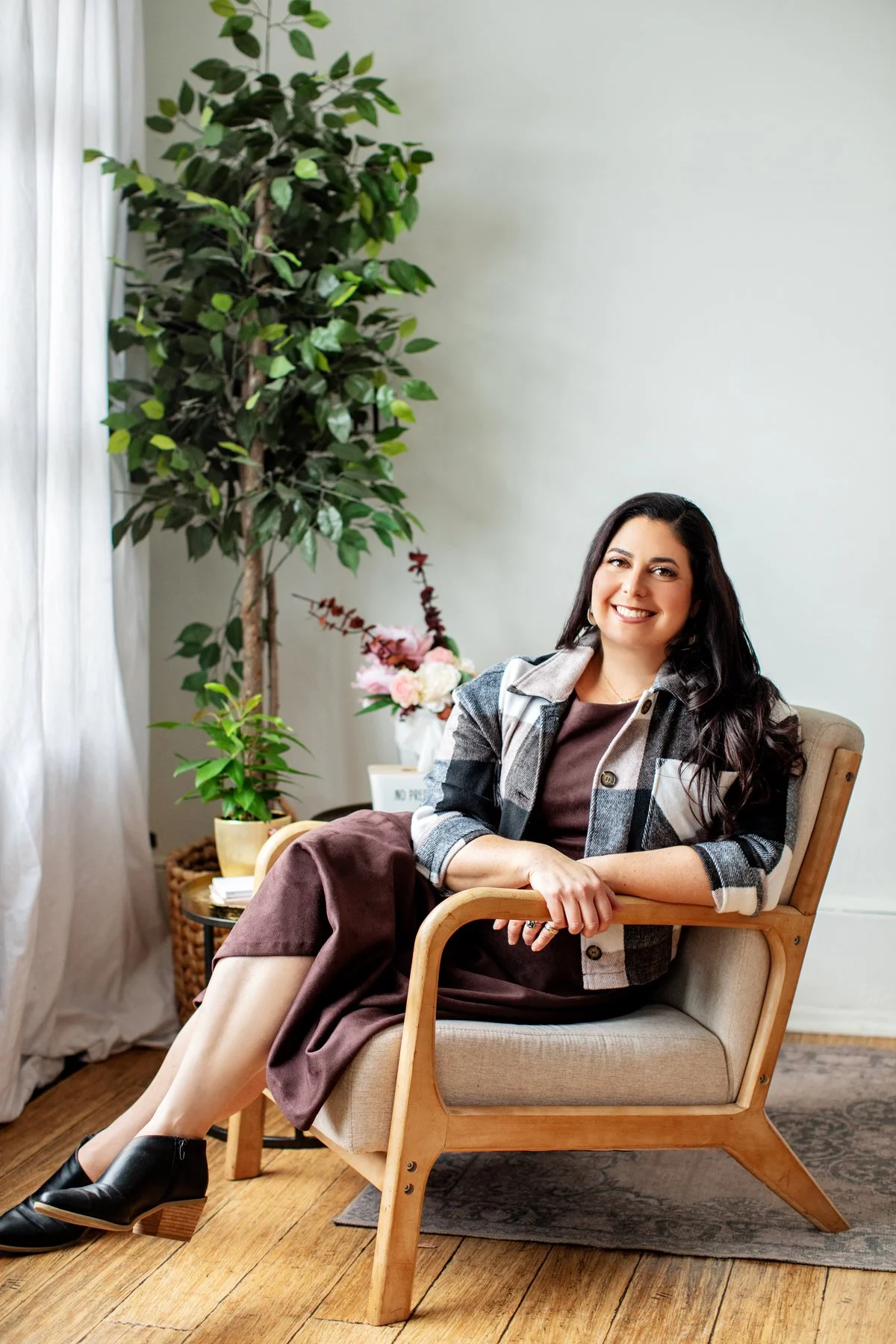 A woman with dark, wavy hair smiling, sitting on a beige armchair with wooden armrests, in a bright room with green plants and flowers in the background.