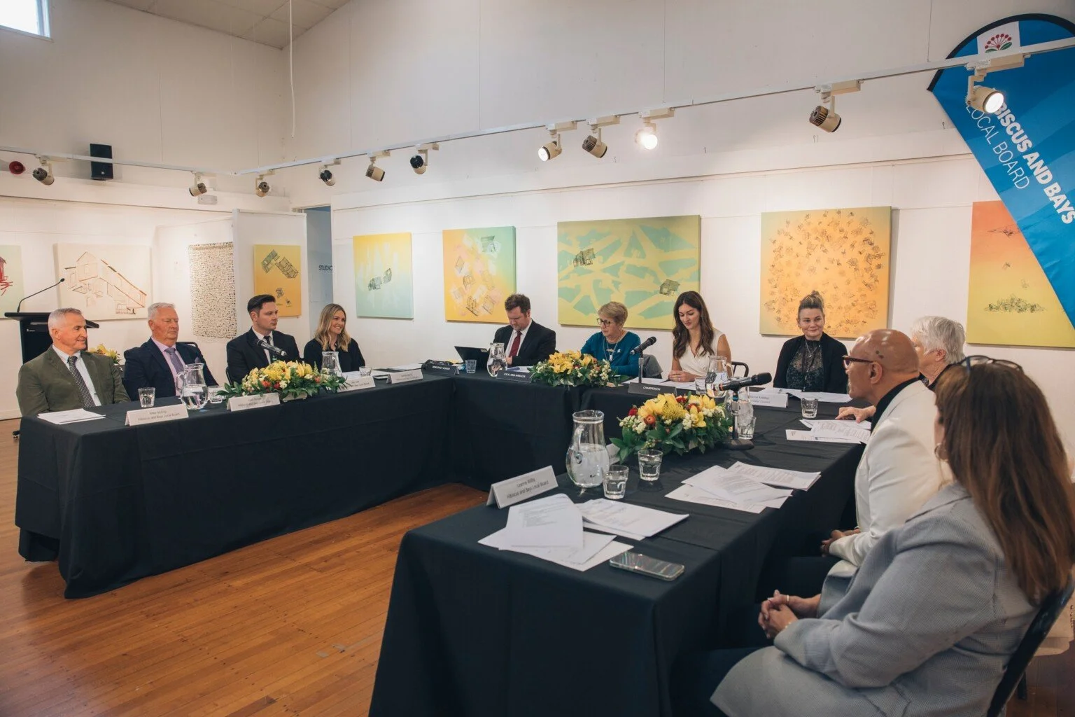Panel discussion or meeting with multiple people seated at a long black table, floral arrangements, and microphones in a gallery with colorful artwork on the walls and a blue banner in the corner.