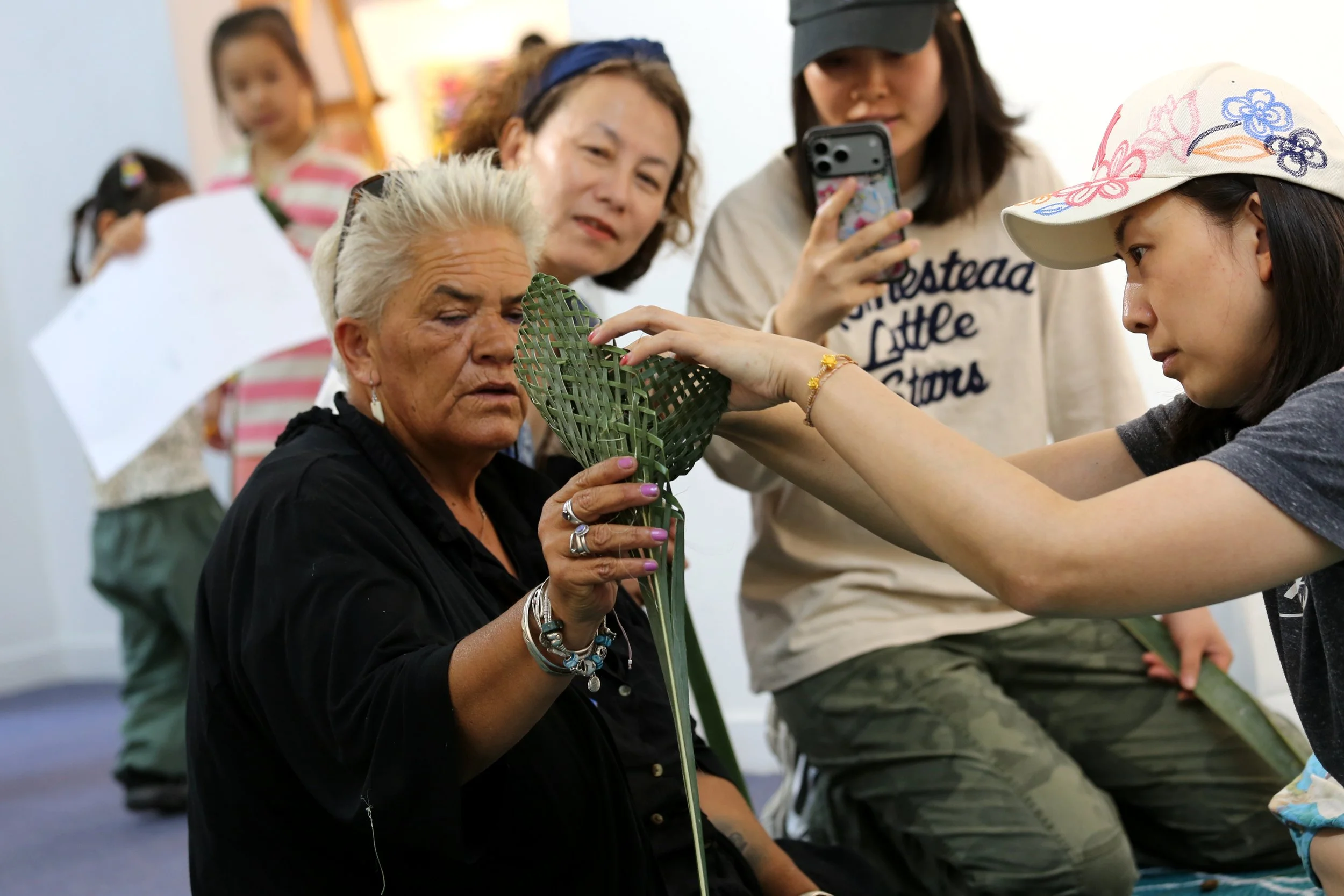 A woman with white hair and numerous rings and bracelets is holding a green, woven palm leaf craft. A young girl is touching the craft and appears to be examining or shaping it. Several other people, including an older woman and a girl in a cap, are observing and taking photos in the background.