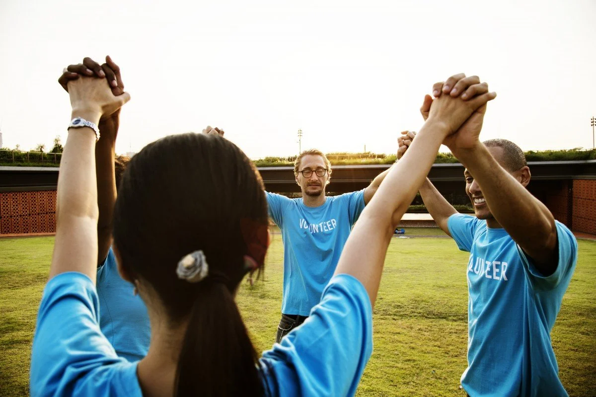 Group of volunteers in blue shirts holding hands in a circle on a grassy field at sunset.