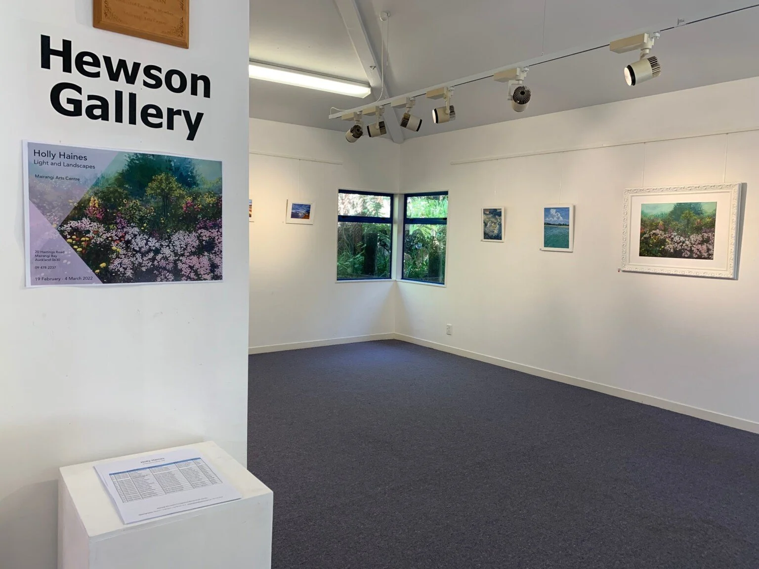 Interior of Hewson Gallery with framed artwork on white walls, spotlight track lighting, and a window showing greenery outside.