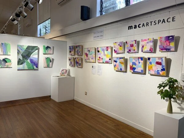 Art gallery displaying abstract paintings with colorful shapes, mounted on white walls, in a space labeled 'macARTSPACE.'