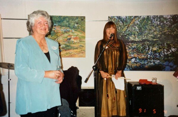 An elderly woman with curly gray hair in a light blue jacket standing and smiling next to a woman with straight brown hair in a gold and black dress speaking into a microphone at an art gallery or event. Two landscape paintings are hung on the wall behind them.