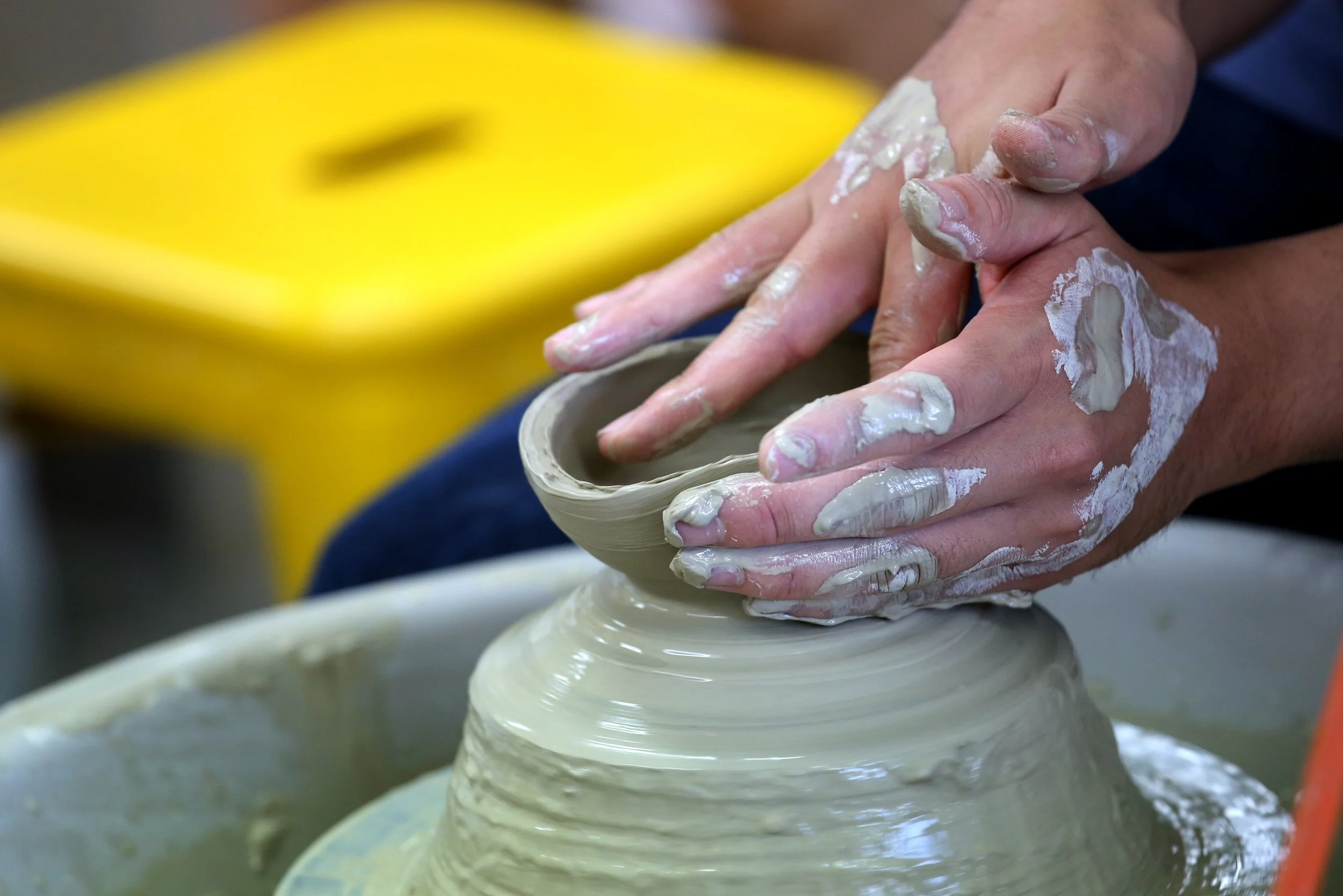 Hands shaping a clay pottery wheel, with wet clay on fingers, in a pottery studio.