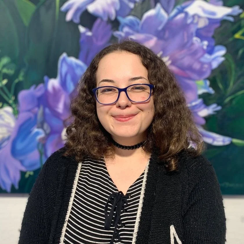 A young woman with curly brown hair, wearing glasses, a black choker, and a black and white striped top, standing in front of a large purple and blue floral painting.