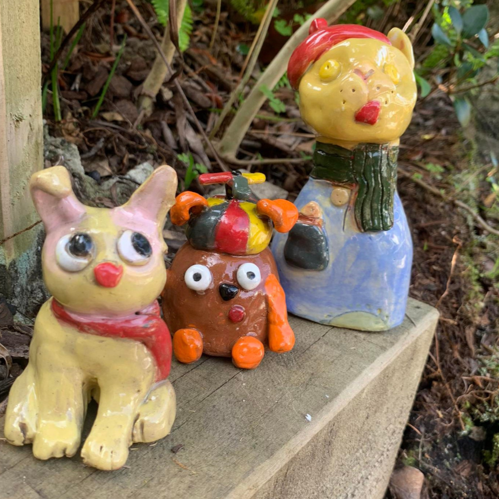 Three colorful ceramic figurines of animals are placed on a wooden surface outdoors. The figurines include a yellow cat with a red bow, a brown dog with a multicolored hat, and a yellow bear with a green scarf and red hat, set against a background of soil and plants.