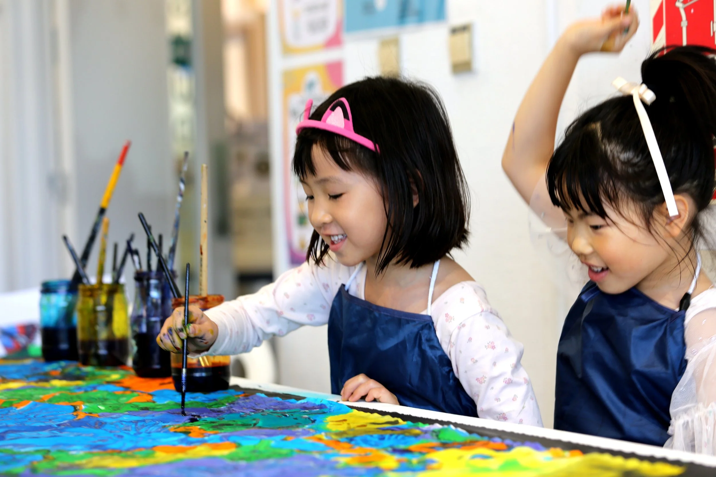 Two young girls painting on a large canvas with colorful paints in a classroom setting.