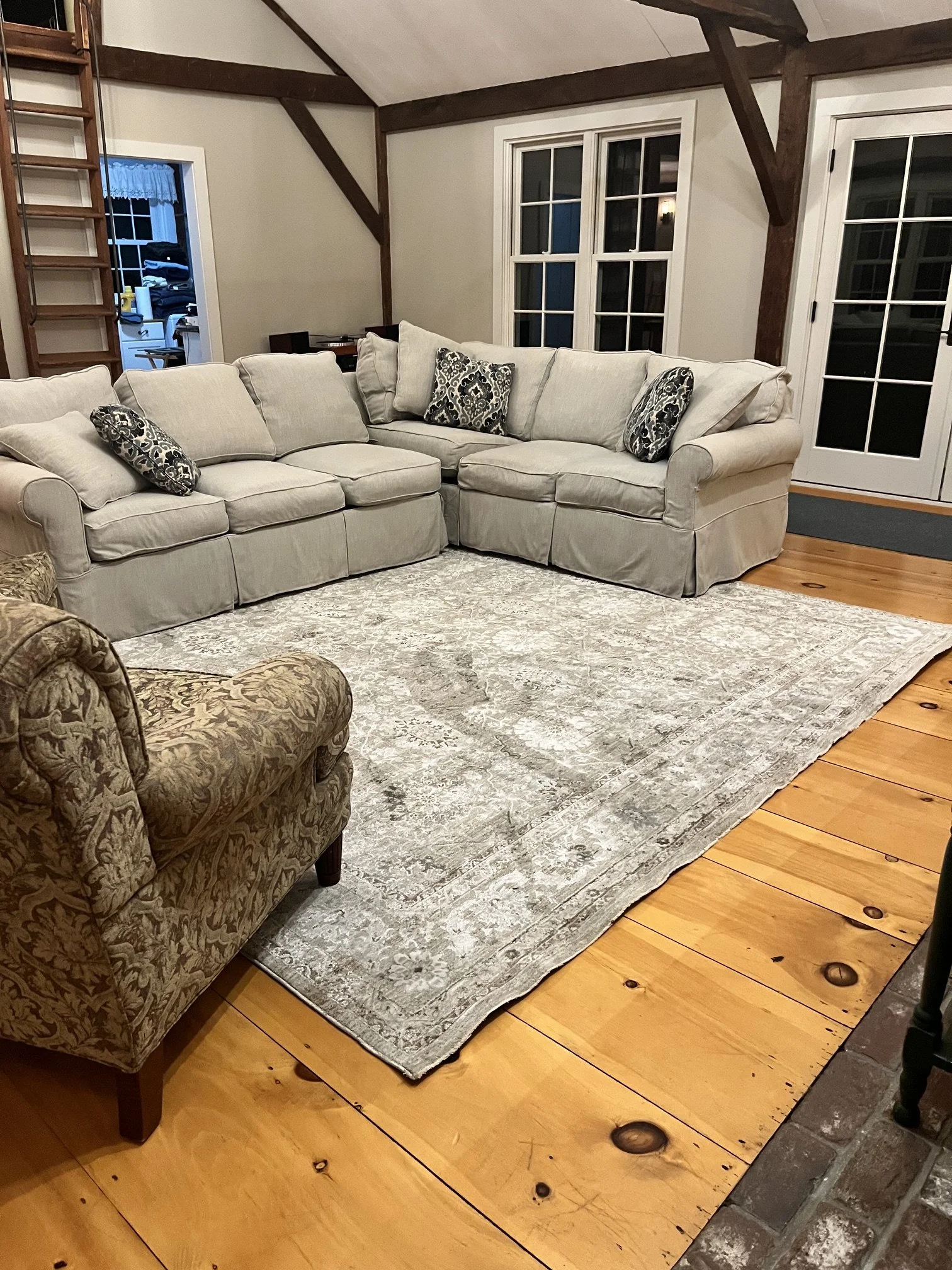 A living room with a beige sofa, patterned throw pillows, and a patterned area rug. There are large windows, wooden floors, and exposed ceiling beams.