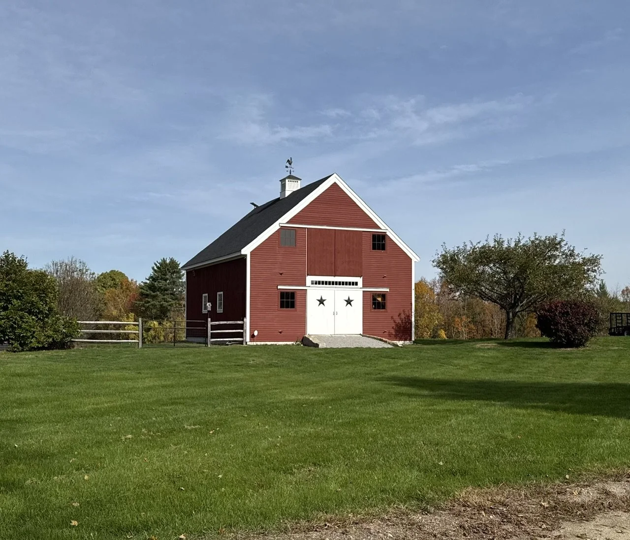 Red barn with white trim and star decorations on the doors, surrounded by green grass and trees, under a blue sky.