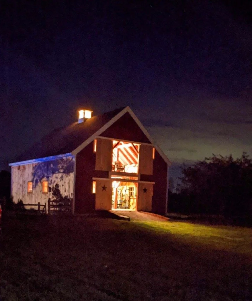 A barn-like structure illuminated from inside at night, with its large door open, revealing a decorated interior. The barn has a steep roof, a small cupola, and is set on a grassy area with trees in the background.