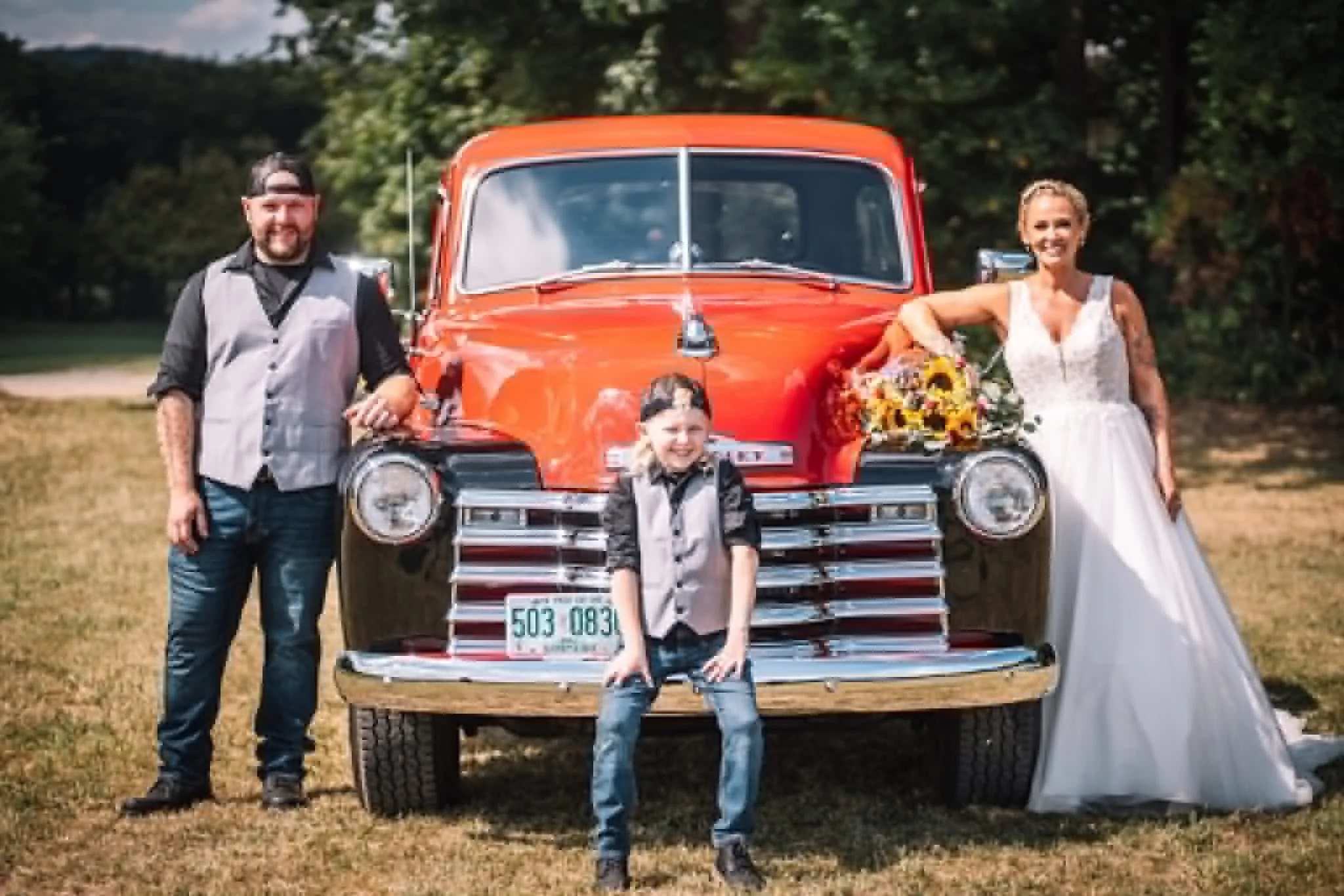 A family of four standing in front of an orange vintage car with a trailer. The father is on the left wearing a plaid vest and jeans, the mother on the right in a white dress holding a bouquet, a girl in a gray vest and jeans stands in front of the car, and a boy in a plaid shirt and jeans is at the front of the trailer. The scene is outdoors with trees and grass.