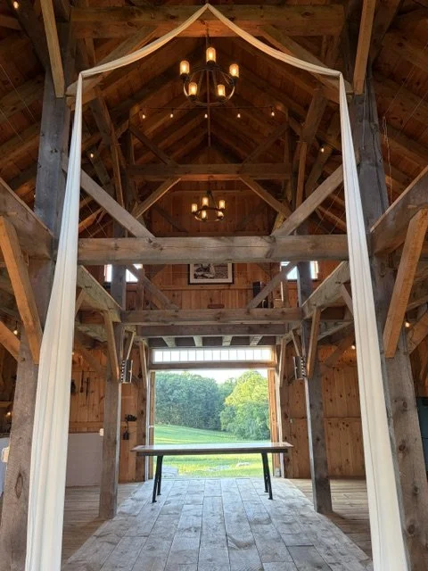 Interior of a rustic wooden barn with a high ceiling, exposed beams, and chandeliers. There is a large open garage-style door with a view of green trees and grass outside. A table is positioned in front of the door.