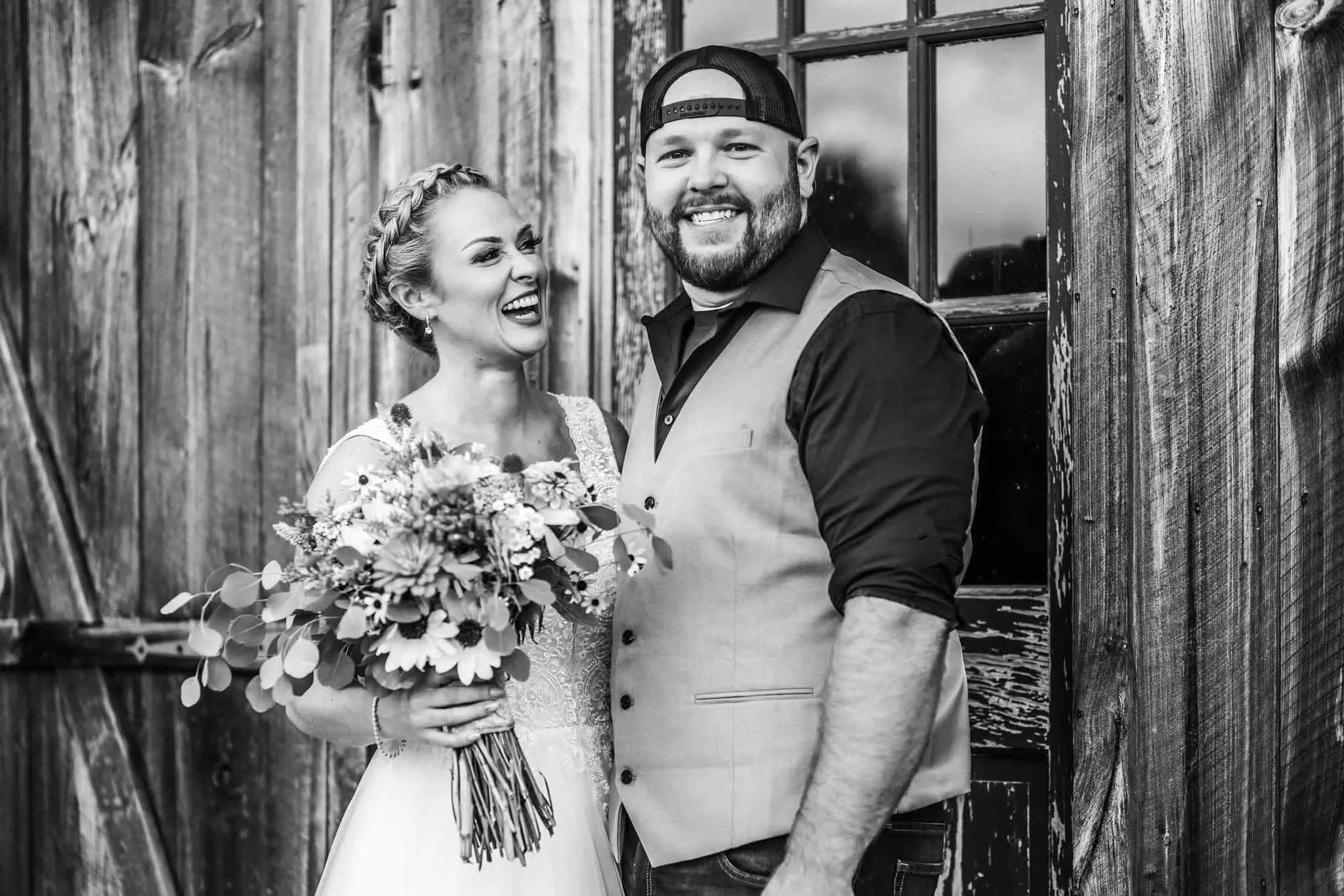 A smiling woman in a wedding dress holding a bouquet stands next to a man in a vest and baseball cap, both leaning against a rustic wooden wall.
