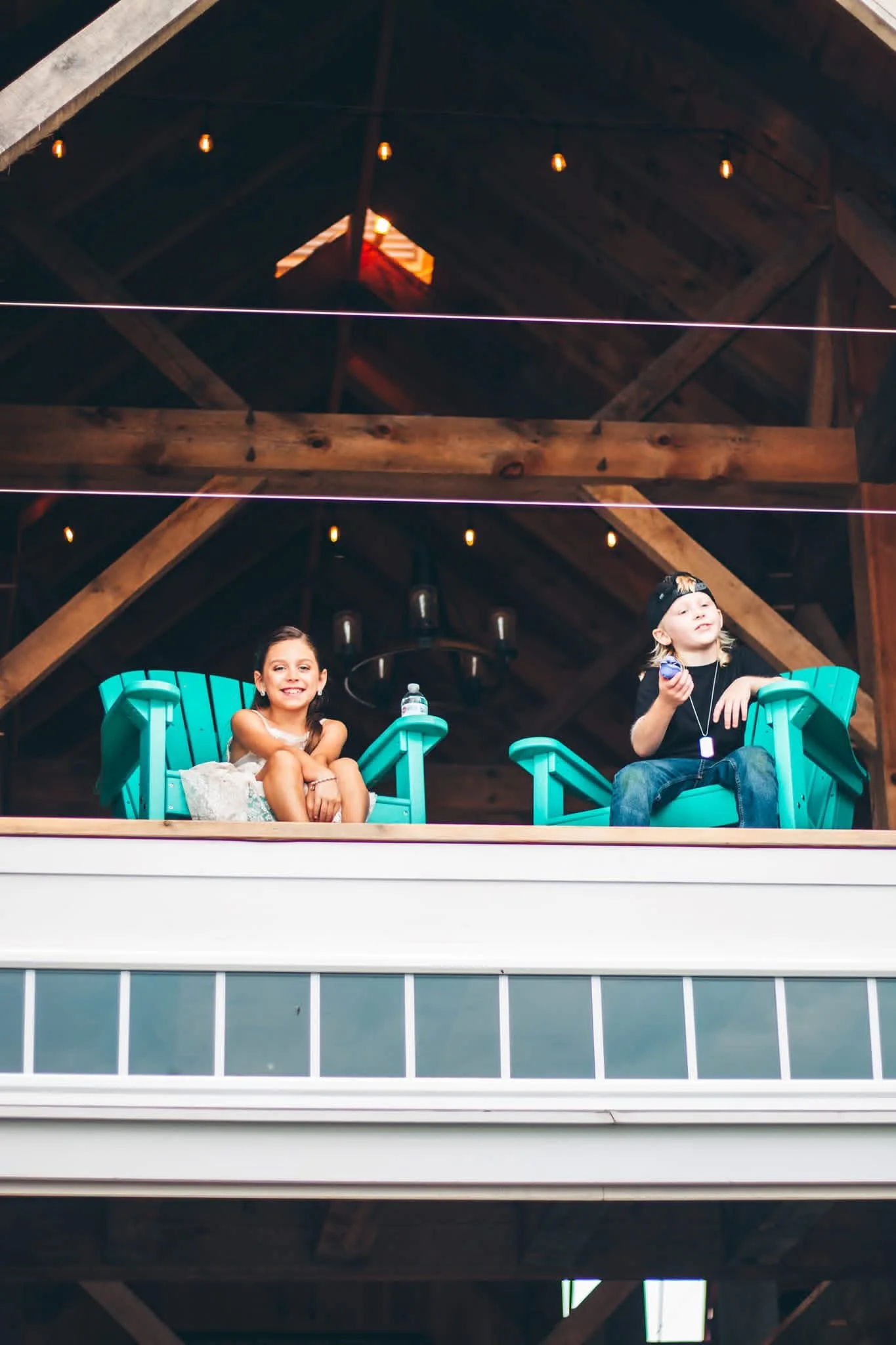 Two children sitting on teal Adirondack chairs on an outdoor balcony, smiling and enjoying the view, with wooden roof beams and string lights above.