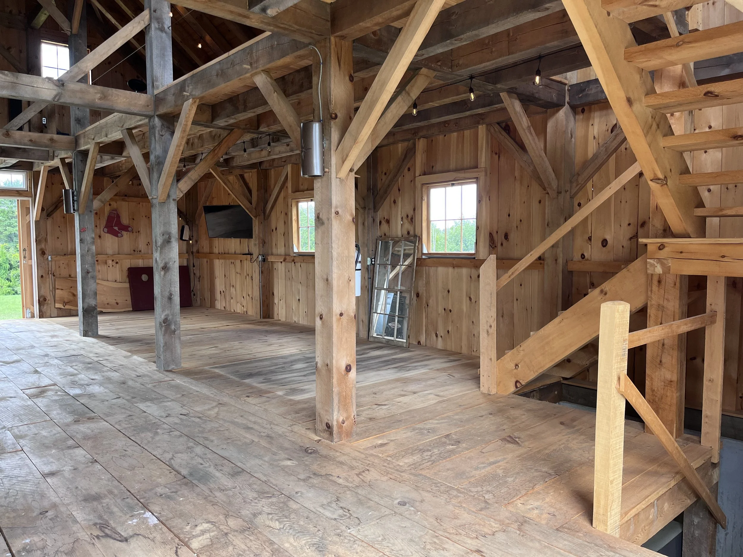 Interior of a wooden house with exposed beams and staircase, with windows providing natural light.