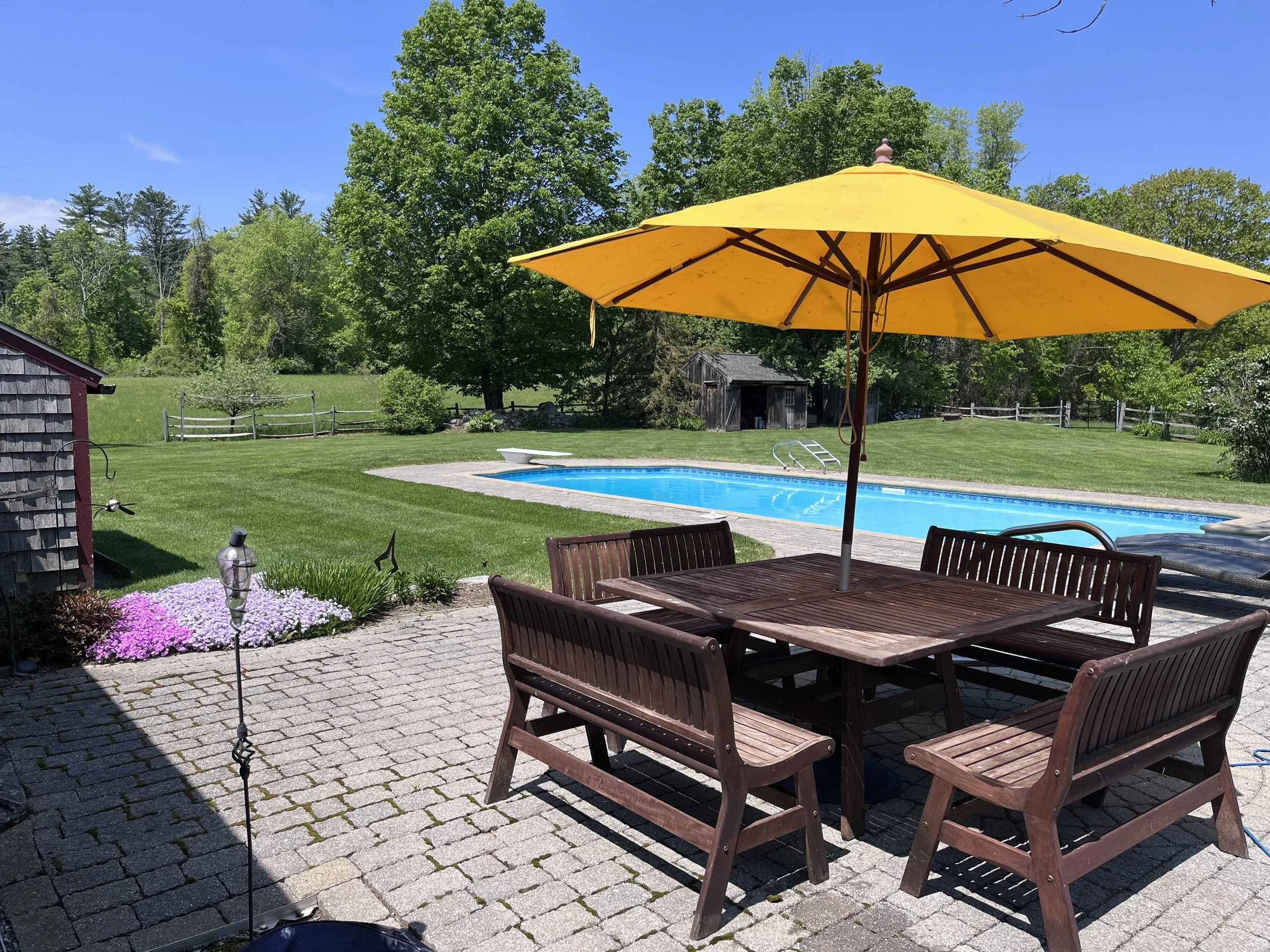A backyard with a swimming pool, outdoor furniture including wooden chairs and a table, a large yellow patio umbrella, green trees, and a clear blue sky.