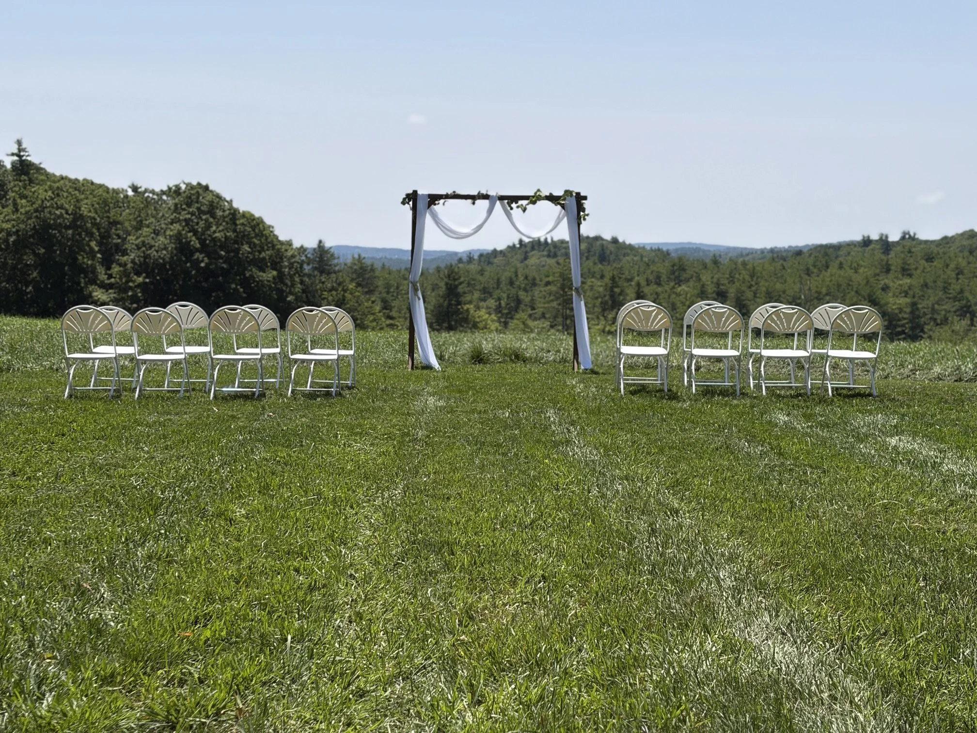 Outdoor wedding ceremony setup with chairs arranged in front of an arch decorated with white fabric and greenery, set on a grassy field with trees and hills in the background under a clear blue sky.