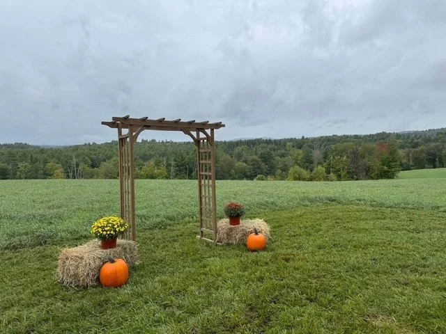 Decorative fall setup with hay bales, pumpkins, potted flowers, and a wooden arch in an open grassy field with trees in the background under a cloudy sky.