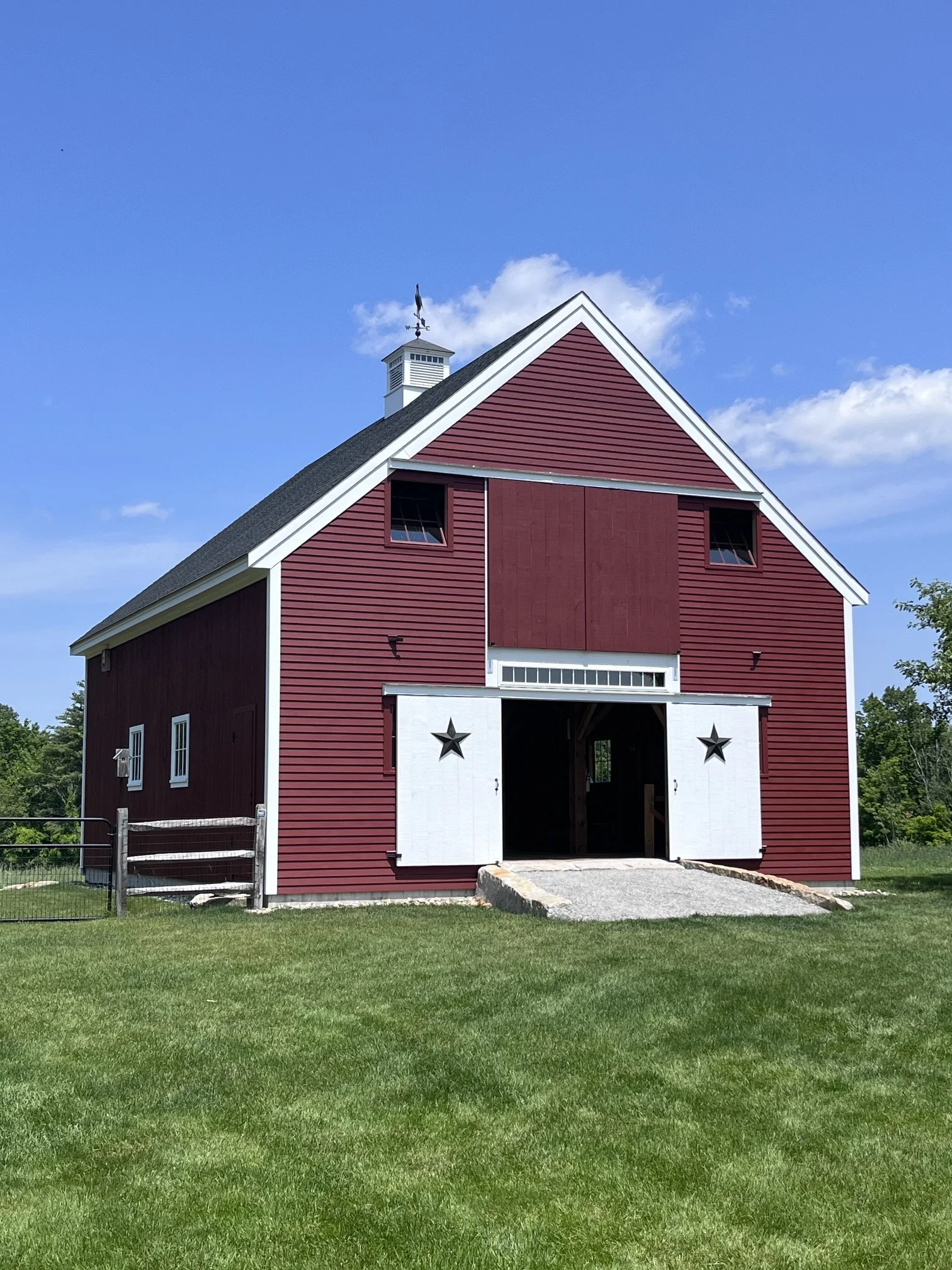 Red barn with white trim, star decoration on white sliding doors, open entrance, small windows, and a weather vane on the roof under blue sky with scattered clouds.