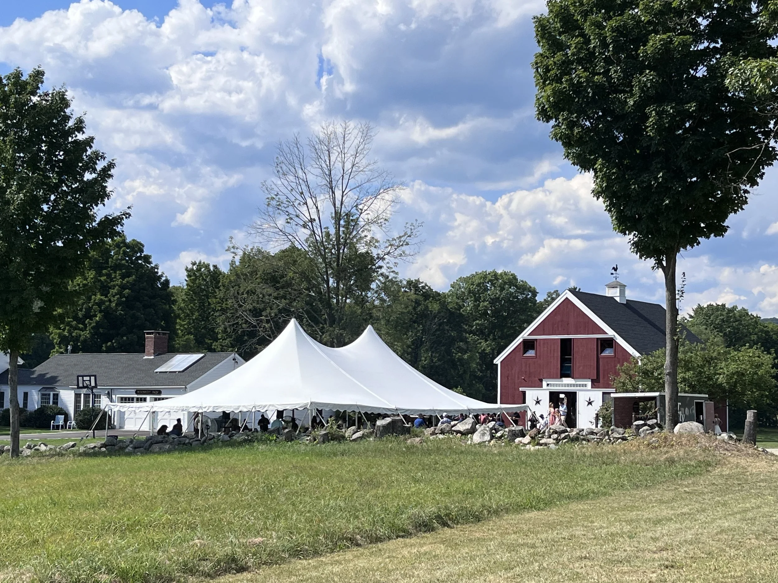 Outdoor scene with white event tents set up on a grassy area, a red barn, trees, and partly cloudy blue sky.