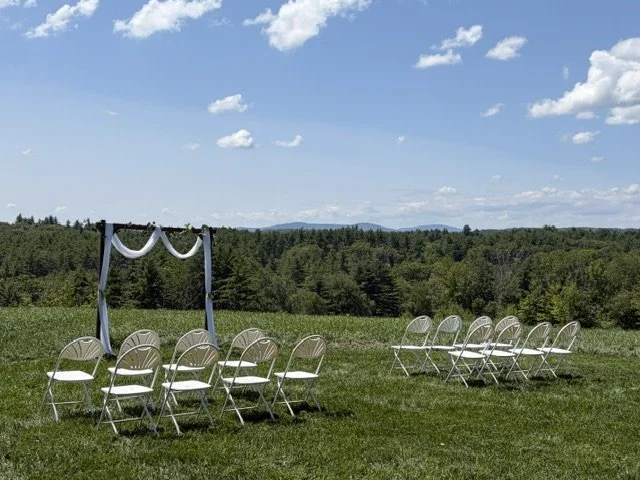 Outdoor wedding ceremony setup with white chairs in front of a grassy field and trees, under a blue sky with scattered clouds.