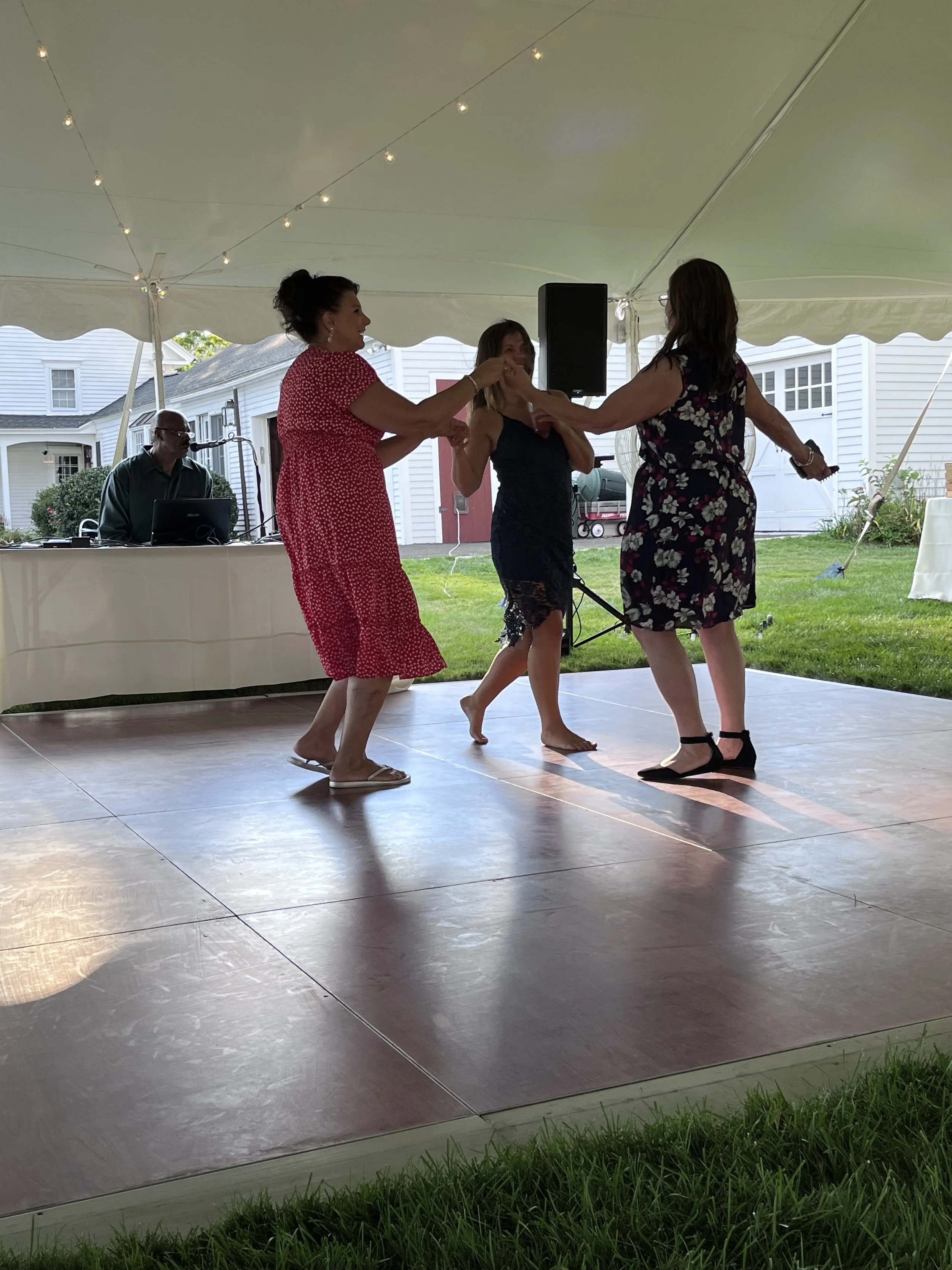 Women dancing under a white canopy at an outdoor event, with a DJ or musician in the background.