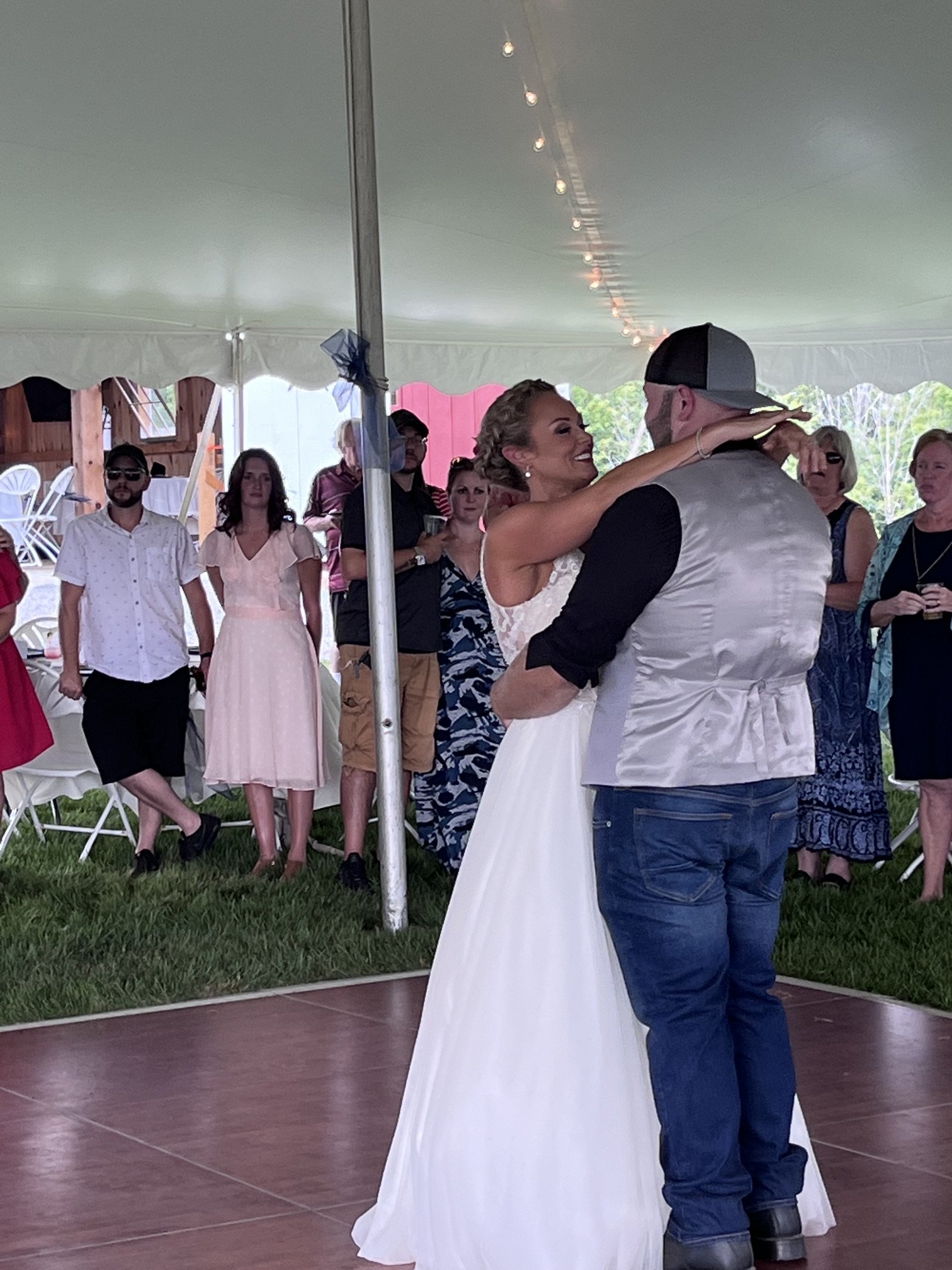 A bride and groom are dancing together under a wedding tent with string lights. Guests watch in the background.