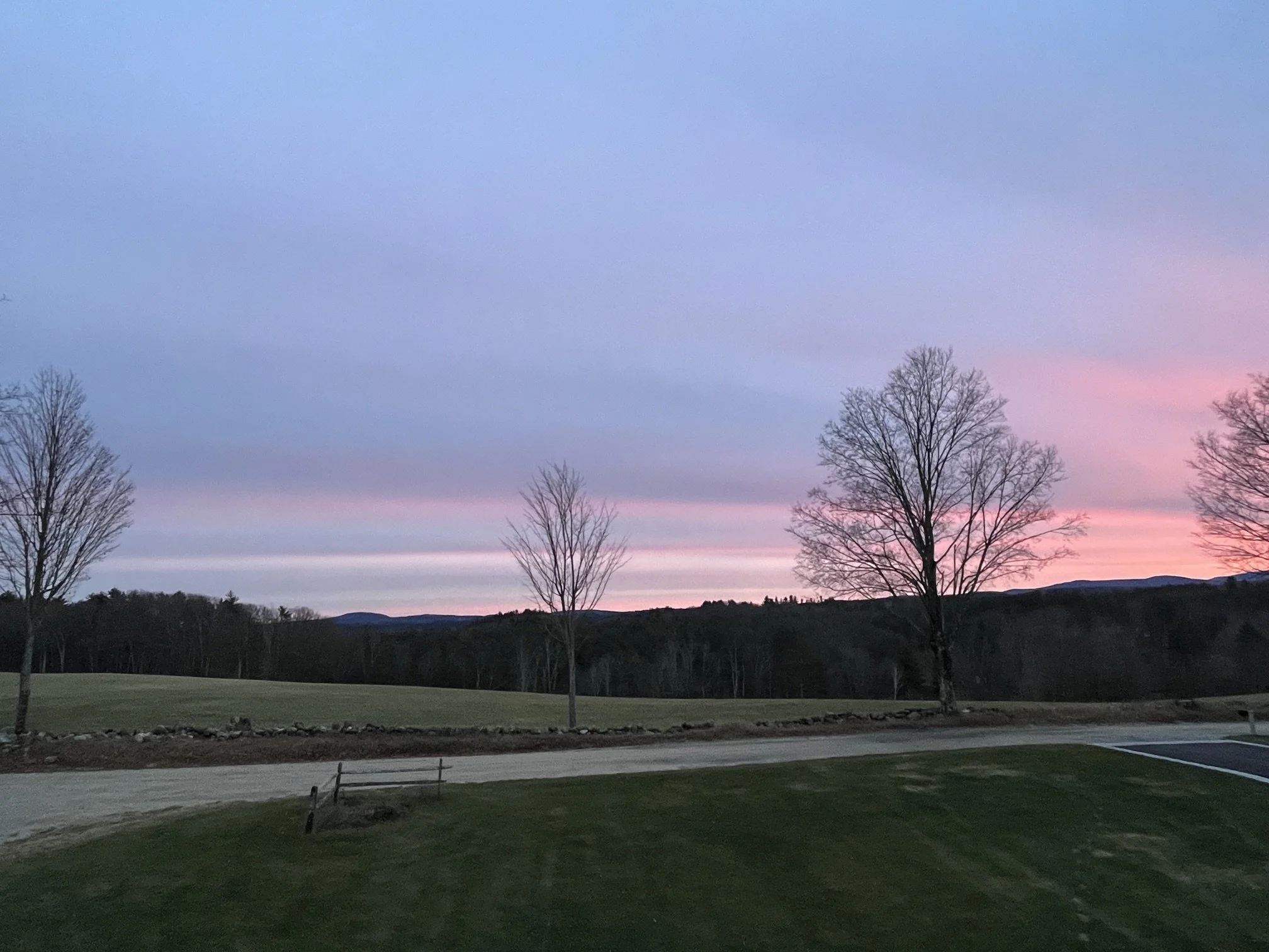 A park scene at sunset with pink and purple hues in the sky, leafless trees, a grassy area, and a parking lot with empty spaces.
