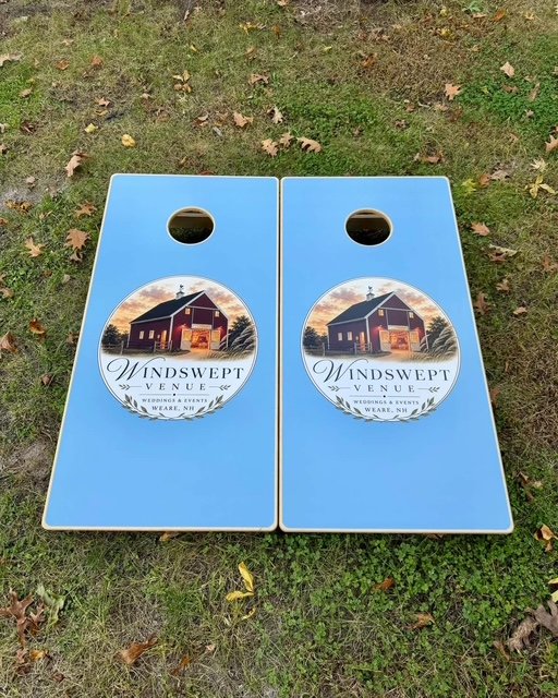 Two cornhole boards with a blue background and a circular logo featuring a barn and the text "Windswept Venue, Wedding Events, Ware, NH," placed on grass with scattered leaves.