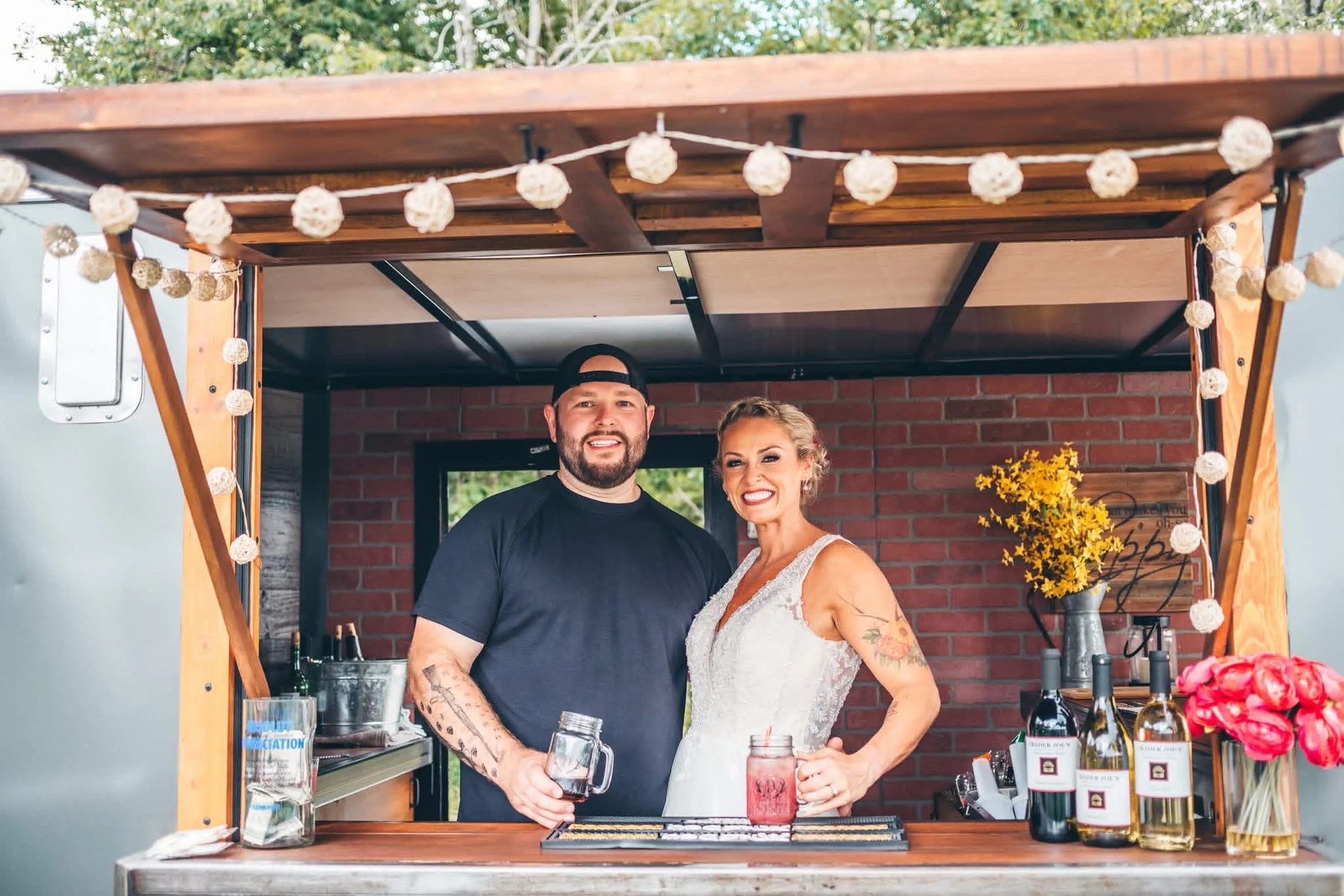 A couple stands behind a bar at an outdoor event, smiling and holding drinks, decorated with string lights and flowers, with a brick wall and trees in the background.