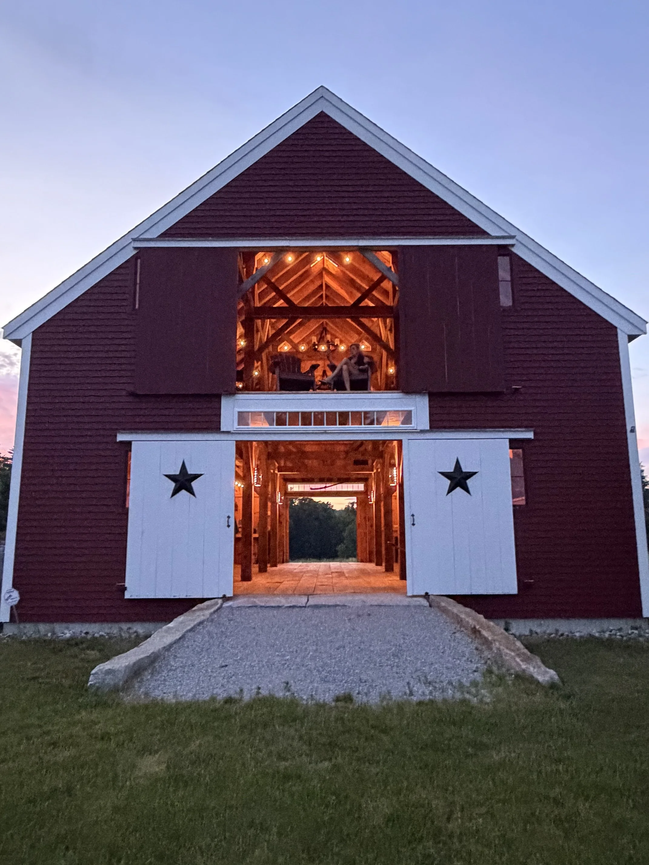 A red barn with white trim and open sliding doors showing a lit interior with string lights, a rocking chair, and a chandelier, in the evening with a grassy area and a gravel pathway in front.