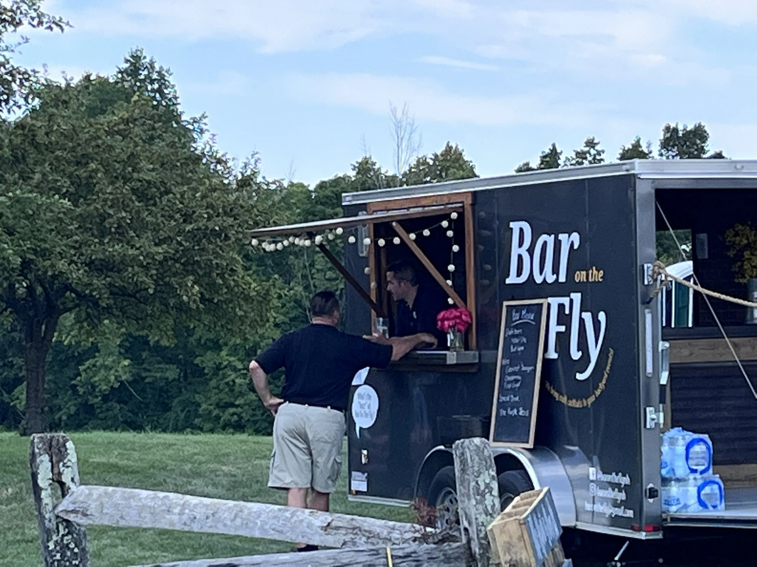 Food truck with a sign advertising a bar on the fly, outdoors on a cloudy day, surrounded by trees, with a woman and a man standing nearby.
