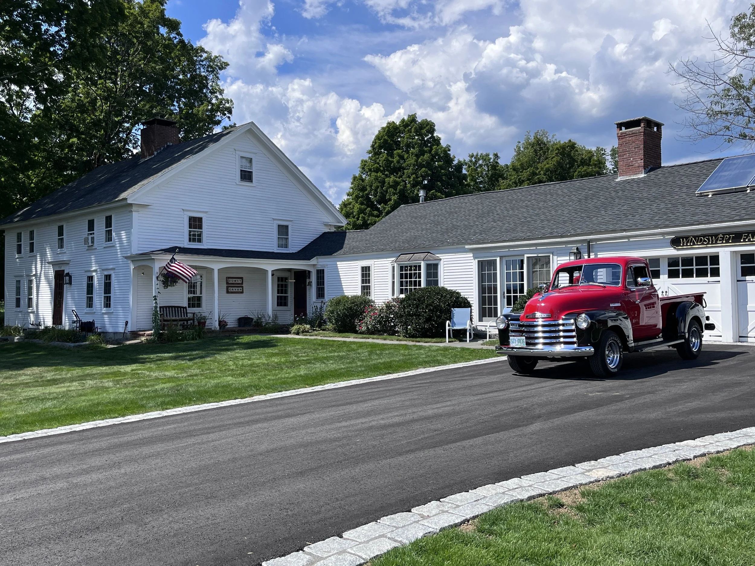 A white two-story house with black roof and brick chimney, front lawn with benches, American flag, and a vintage red truck parked in front, under a partly cloudy sky.