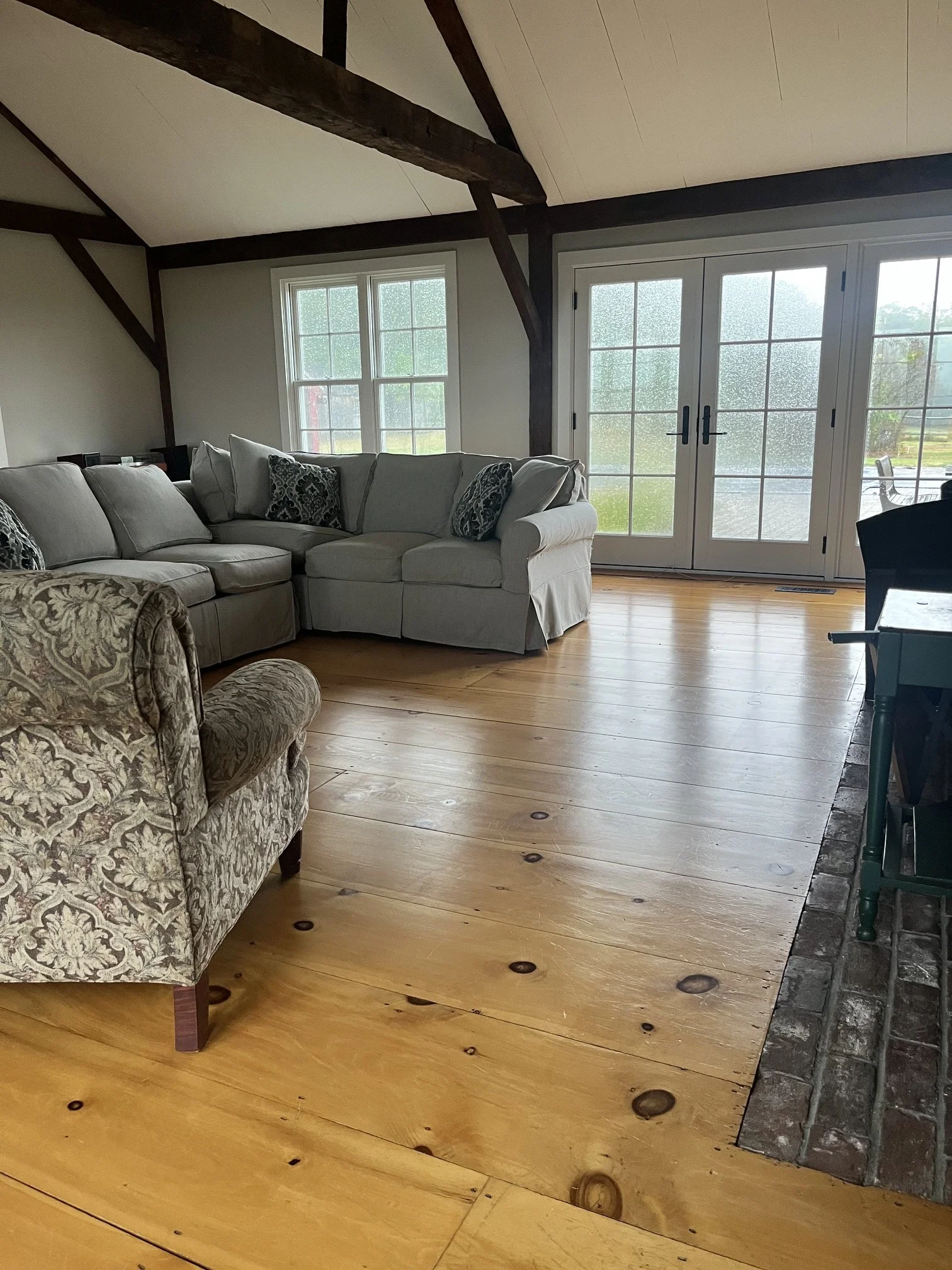 Living room featuring a beige sectional sofa with patterned throw pillows, hardwood flooring, large windows, and glass double doors leading outside. Part of a patterned armchair and a black dining chair are also visible.