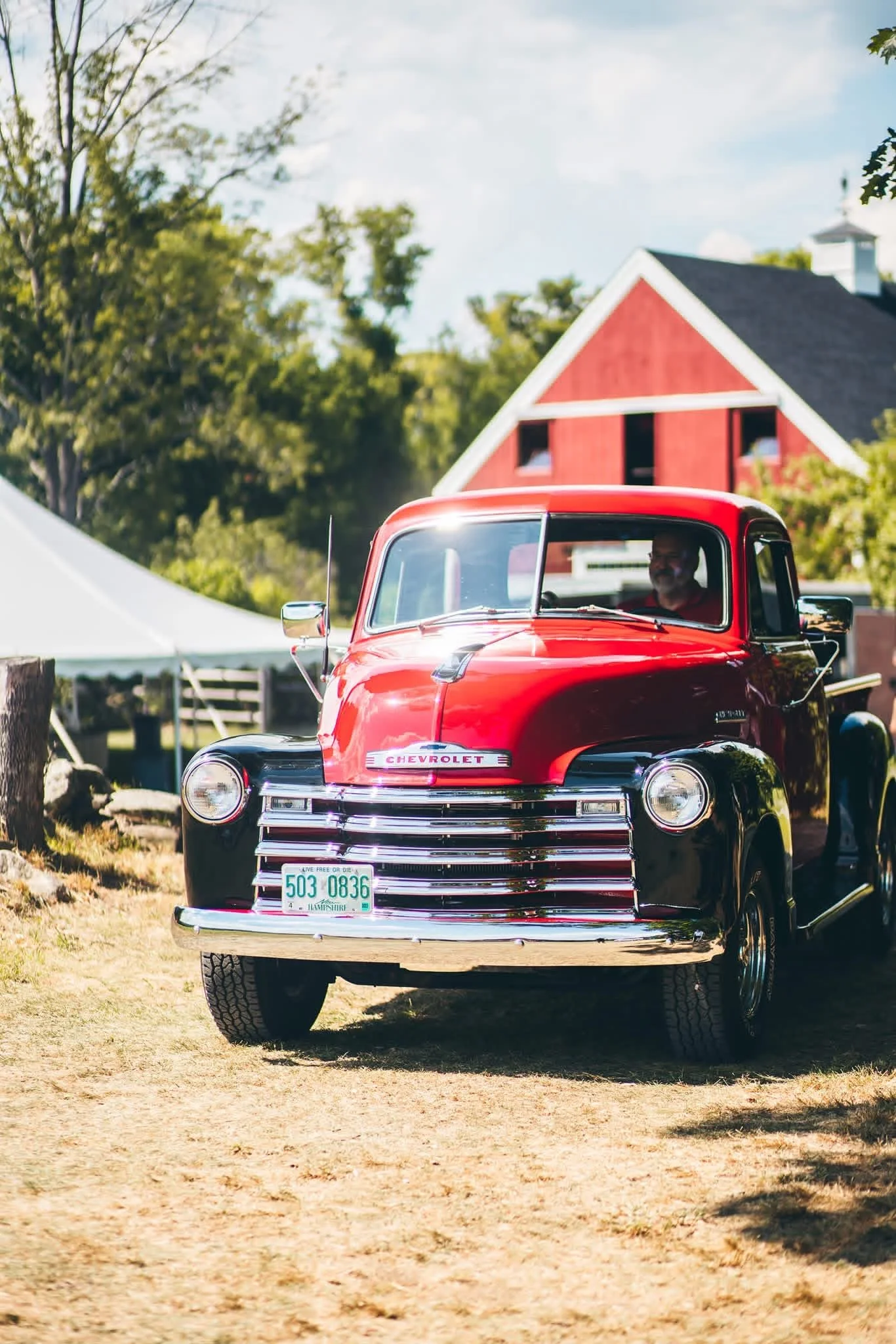 A red vintage Chevrolet truck parked on a dirt ground in front of a barn and trees.