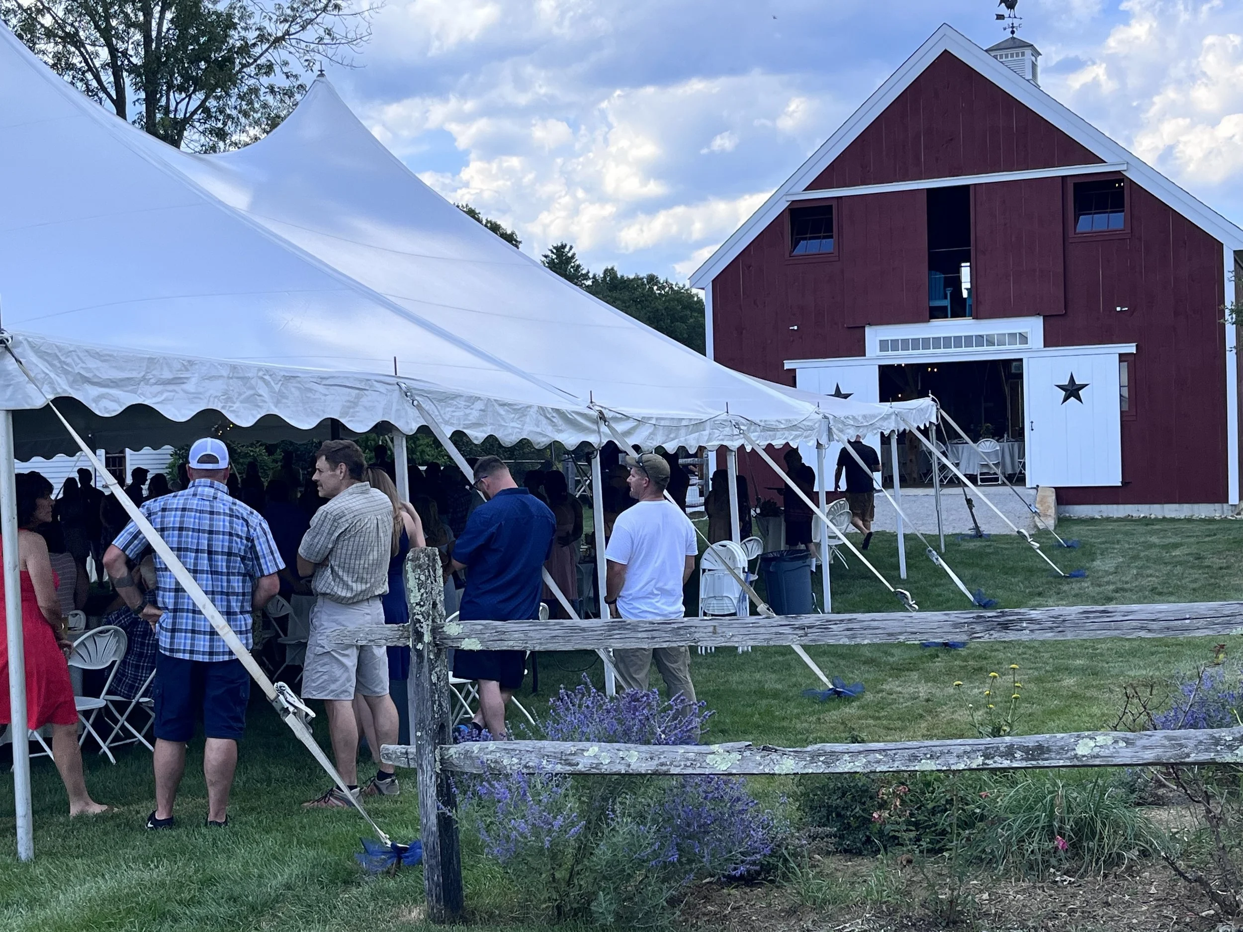 A gathering of people under a large white tent outside a red barn with white trim on a partly cloudy day.