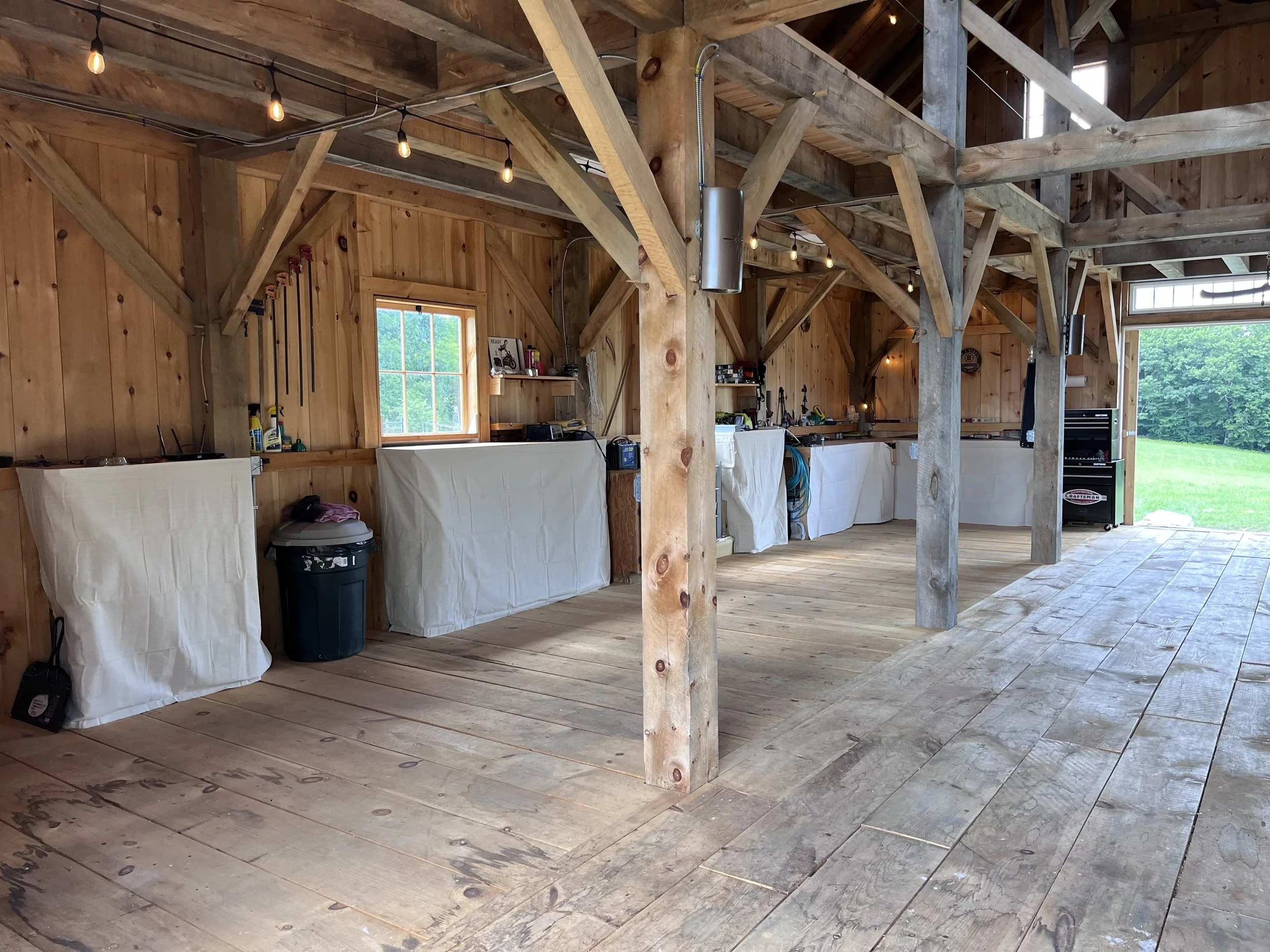 Interior of a wooden building with exposed beams, string lights, and a window showing trees outside. Covered objects and various tools are visible, indicating a workshop or garage setting.