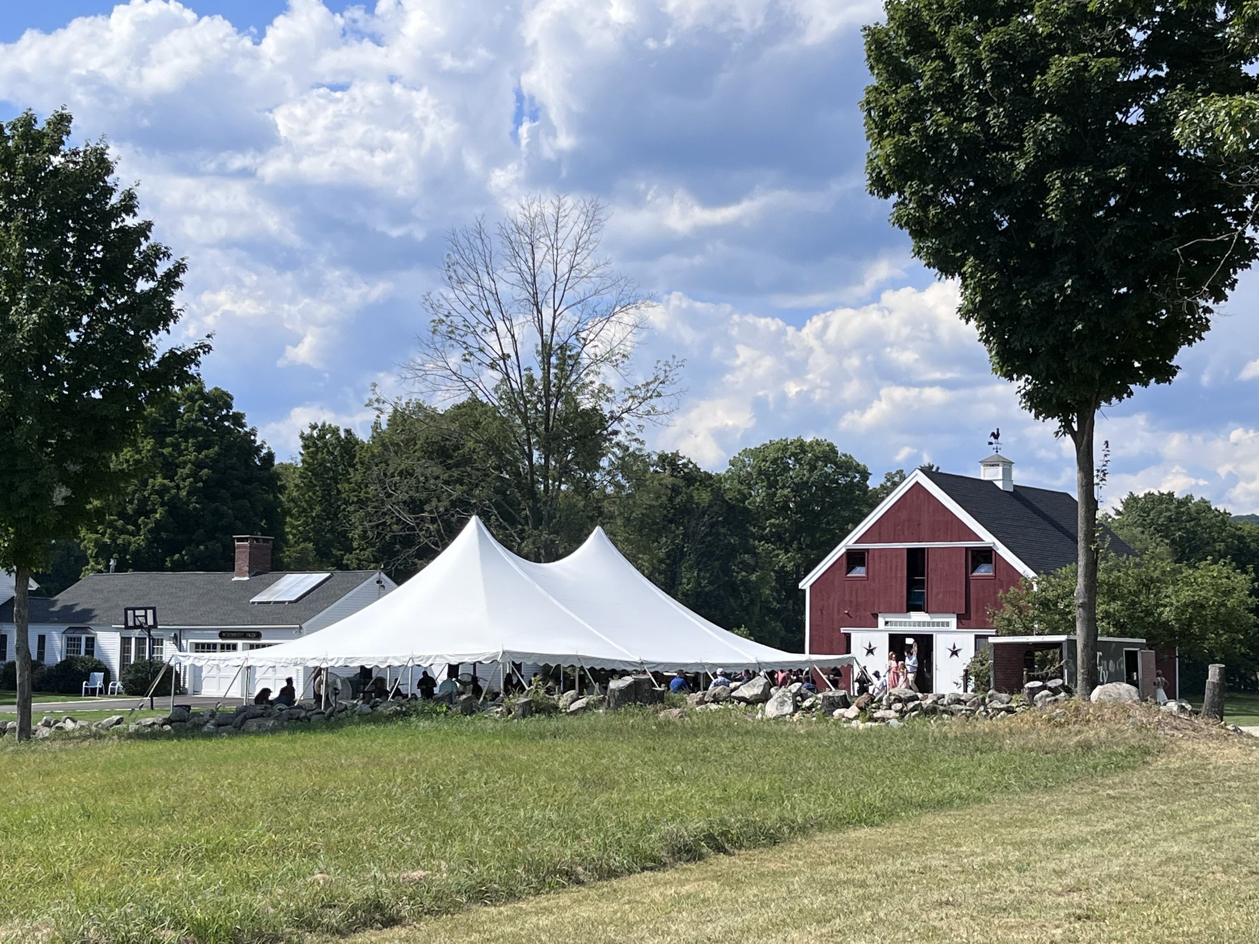 A scene of a outdoor event with a large white tent, people sitting underneath, surrounded by greenery and trees. There are houses in the background with a partly cloudy sky overhead.