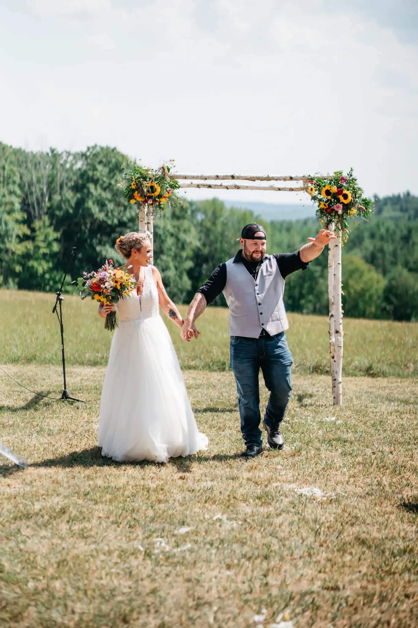 Bride and groom holding hands and walking outdoors during their wedding ceremony, with floral arrangements on a wooden arch in the background.