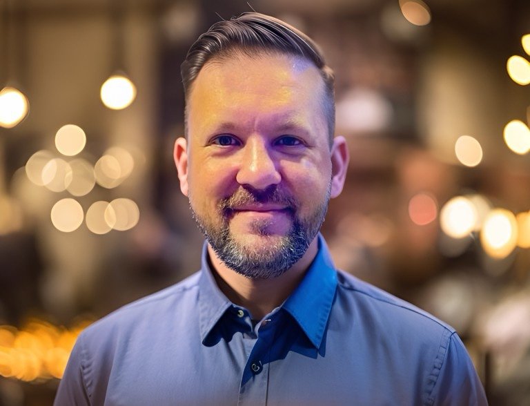 A smiling man with short dark hair, a beard, and wearing a blue collared shirt in a warmly lit setting with bokeh lights in the background.