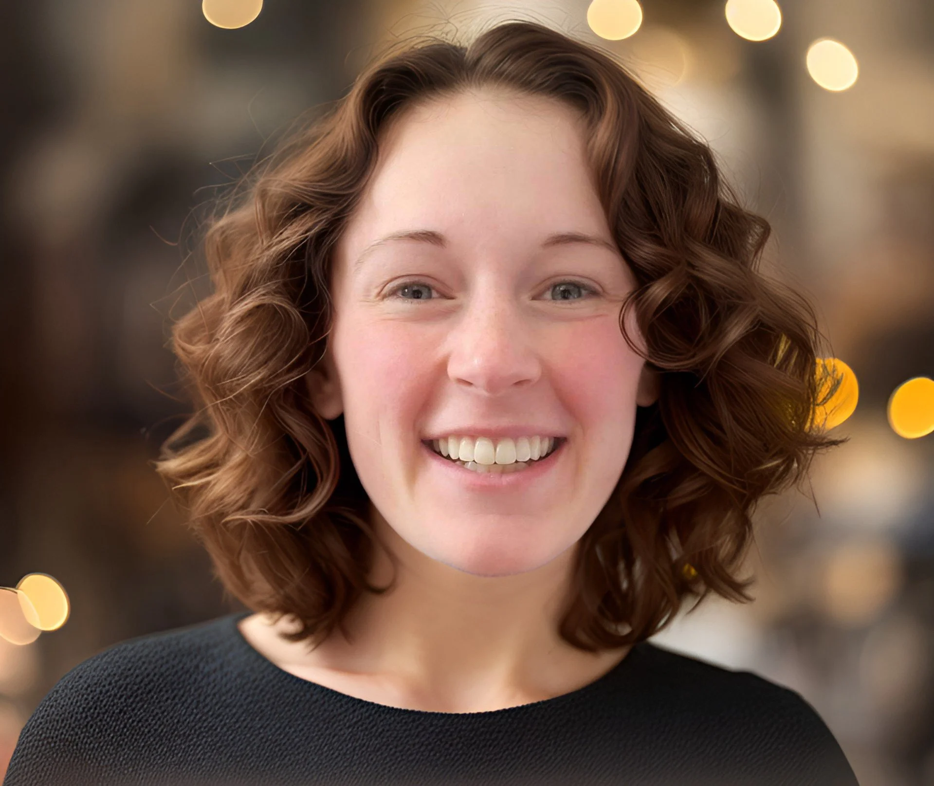 A young woman with curly brown hair smiling in front of a blurred background with bokeh light effects.