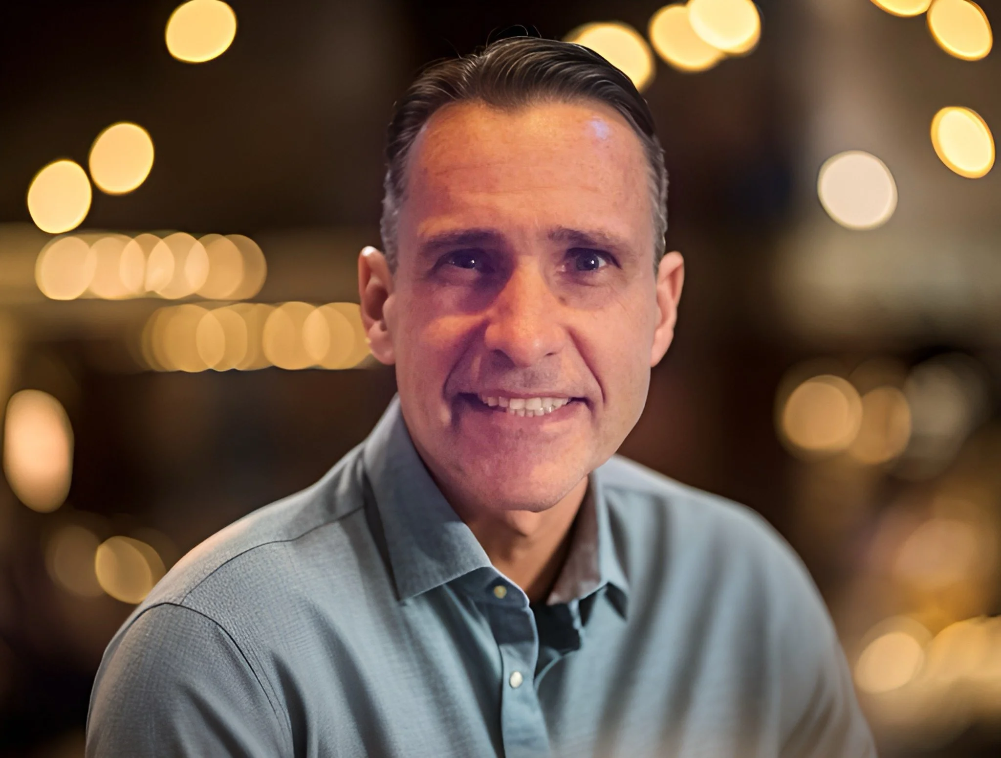 A man with short dark hair wearing a gray collared shirt, smiling with lights blurred in the background.