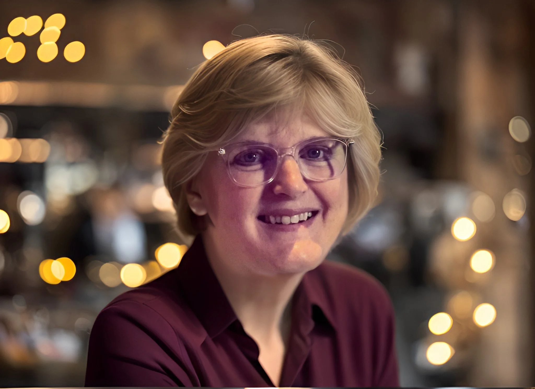 Portrait of a woman with blonde hair and glasses smiling in a warmly lit indoor setting with bokeh lights in the background.