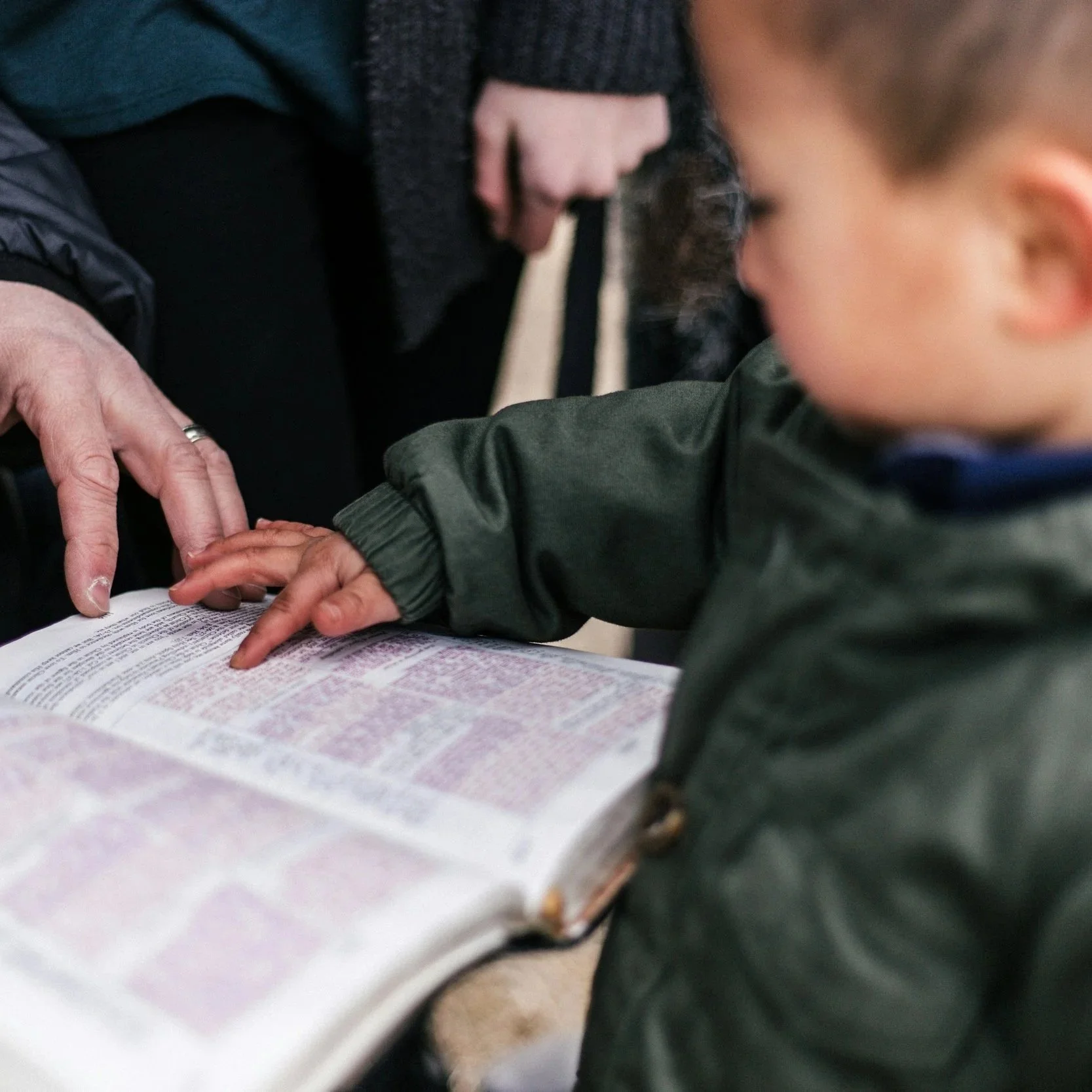 A child touching a Bible or religious book while an adult points to the pages, with a person standing nearby.