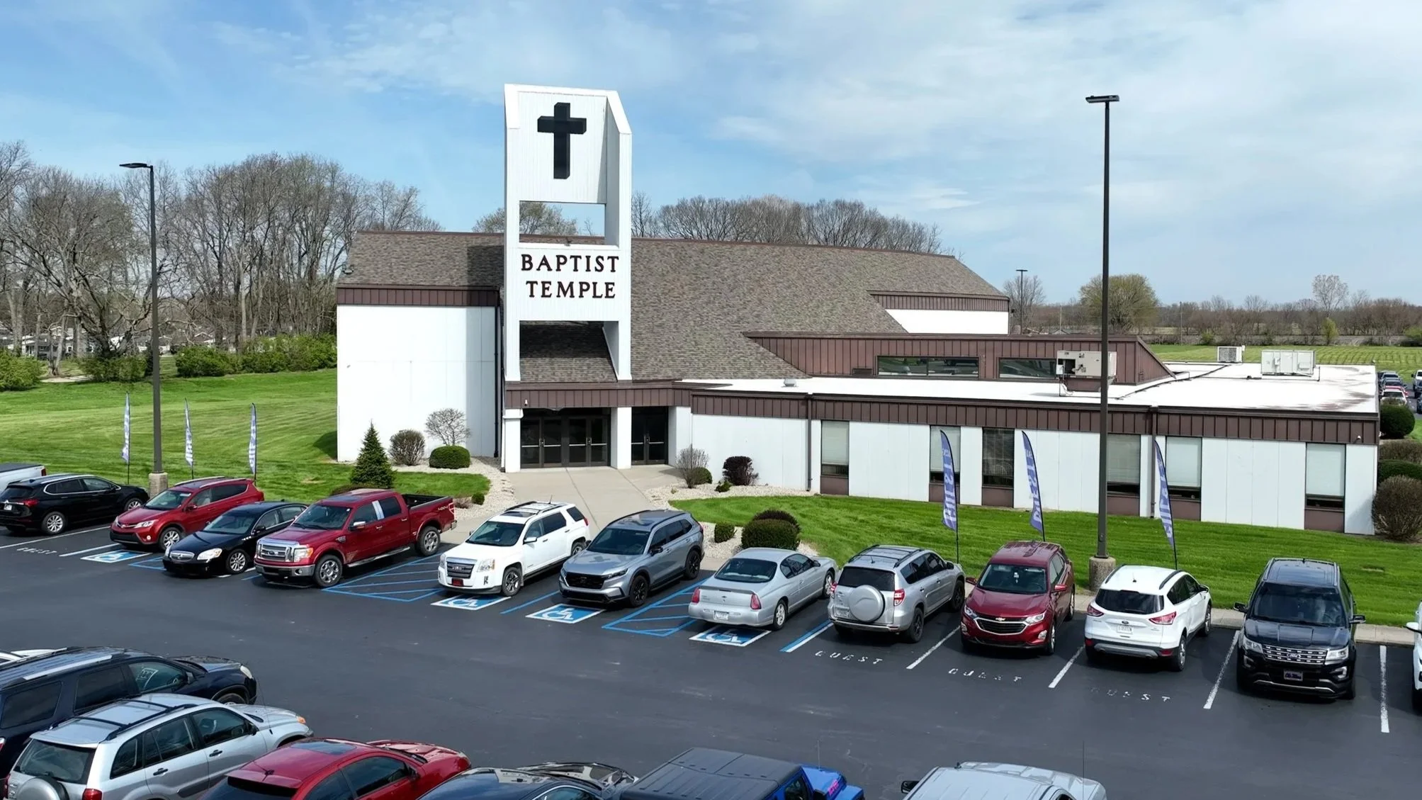 Exterior view of a Baptist Temple church building with a large sign featuring a cross. The parking lot in front has several cars, including designated handicapped spots. Green grass and trees surround the building, under a partly cloudy sky.