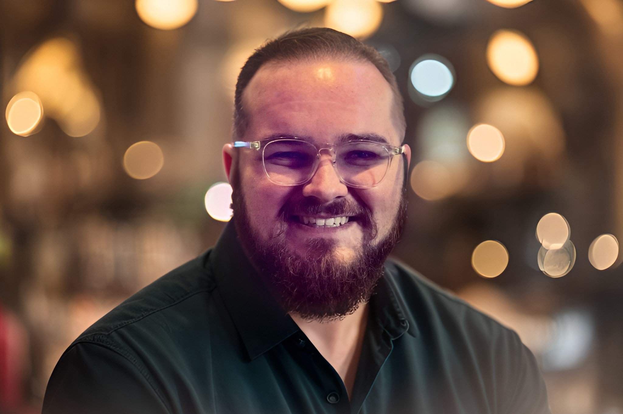 A smiling man with glasses and a beard, wearing a dark shirt, against a blurred background with warm bokeh lights.