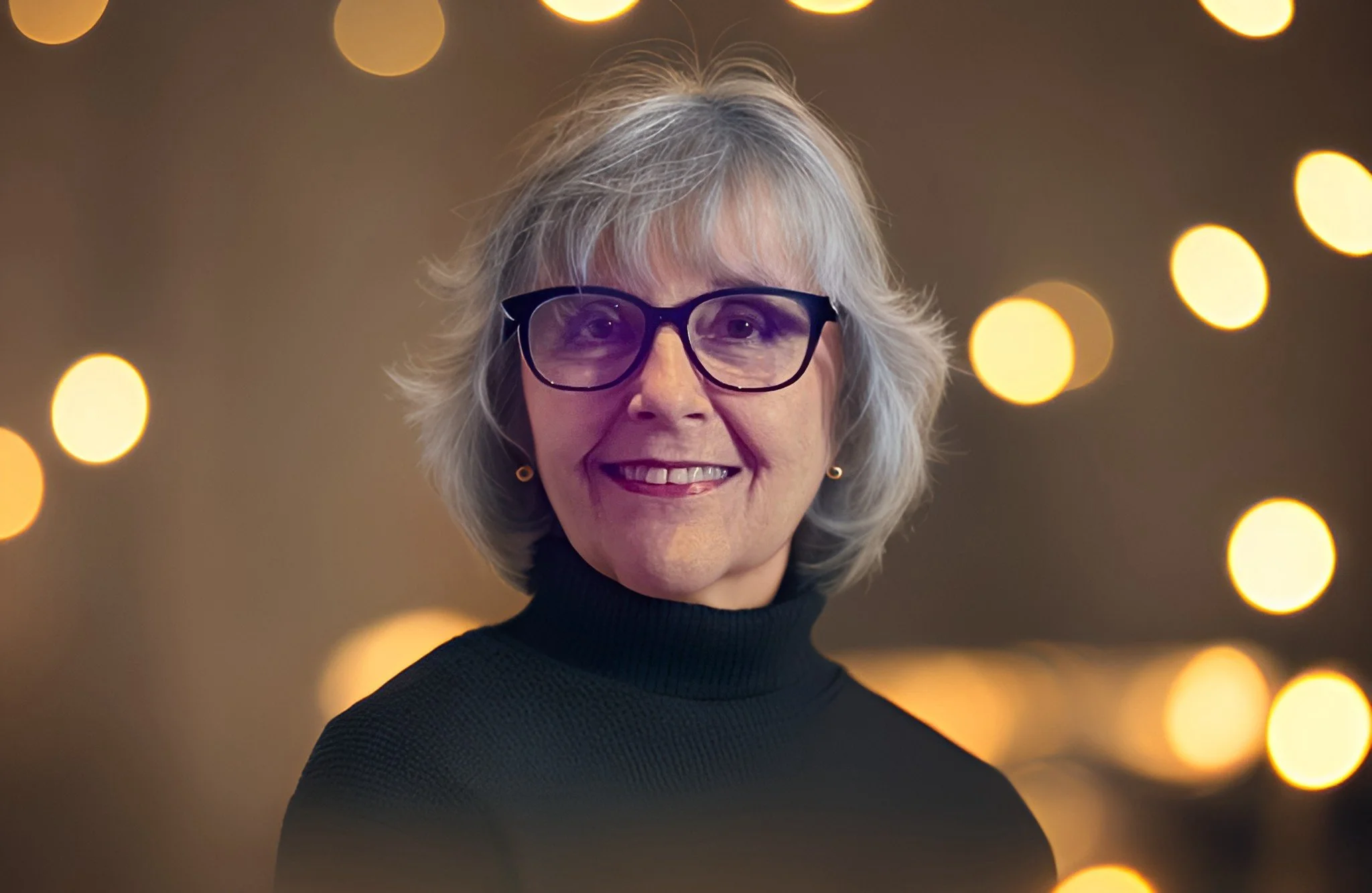 An older woman with gray hair wearing black glasses and a black turtleneck, smiling with a blurred background of warm bokeh lights.